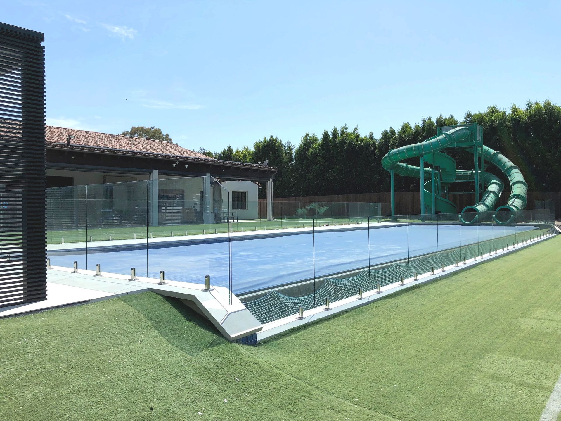 Swimming pool with a water slide, surrounded by a glass fence and green turf. A building is visible in the background.