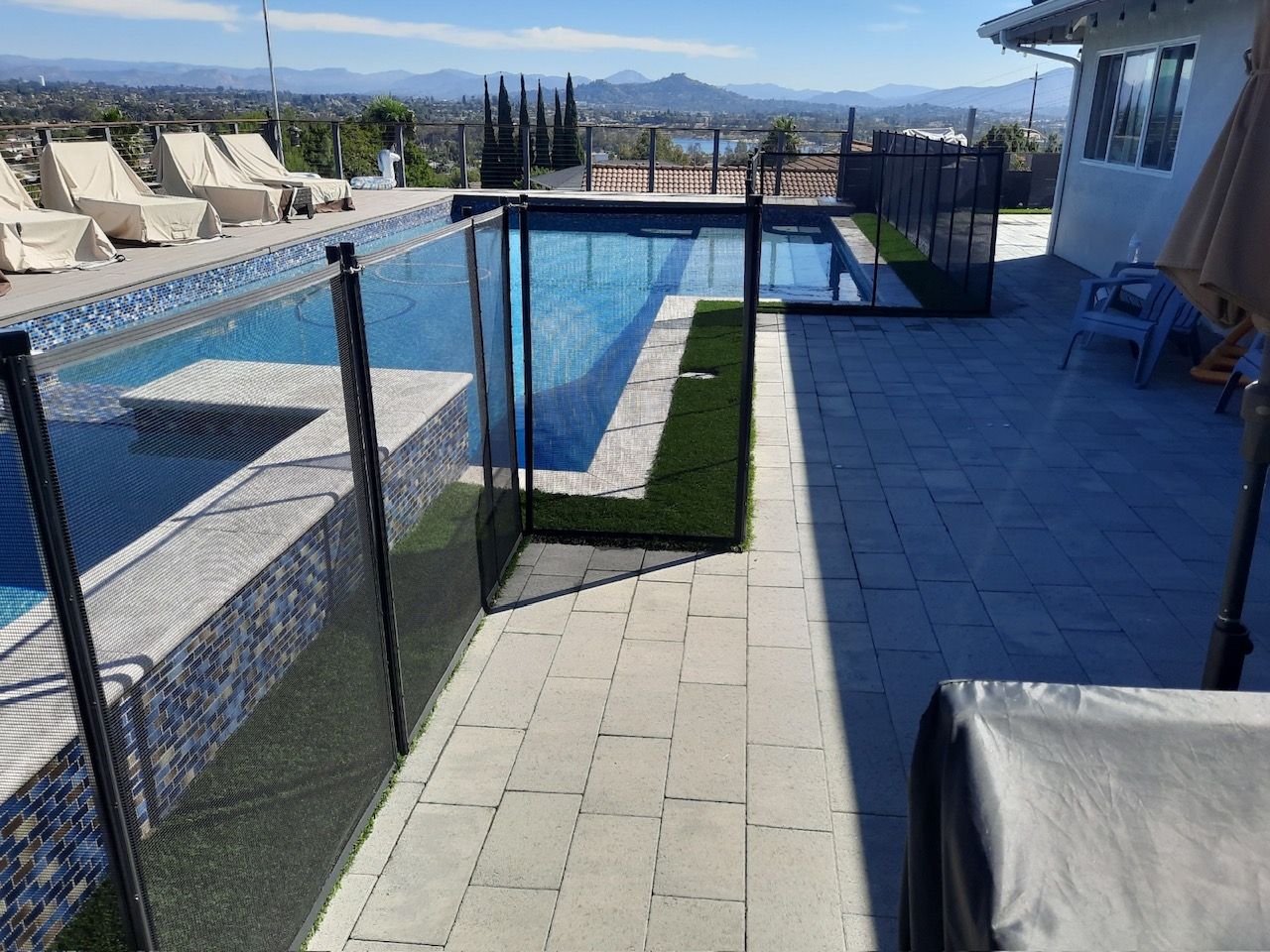 Swimming pool with black mesh safety fence. Paved patio with artificial grass. Sunny outdoor setting.
