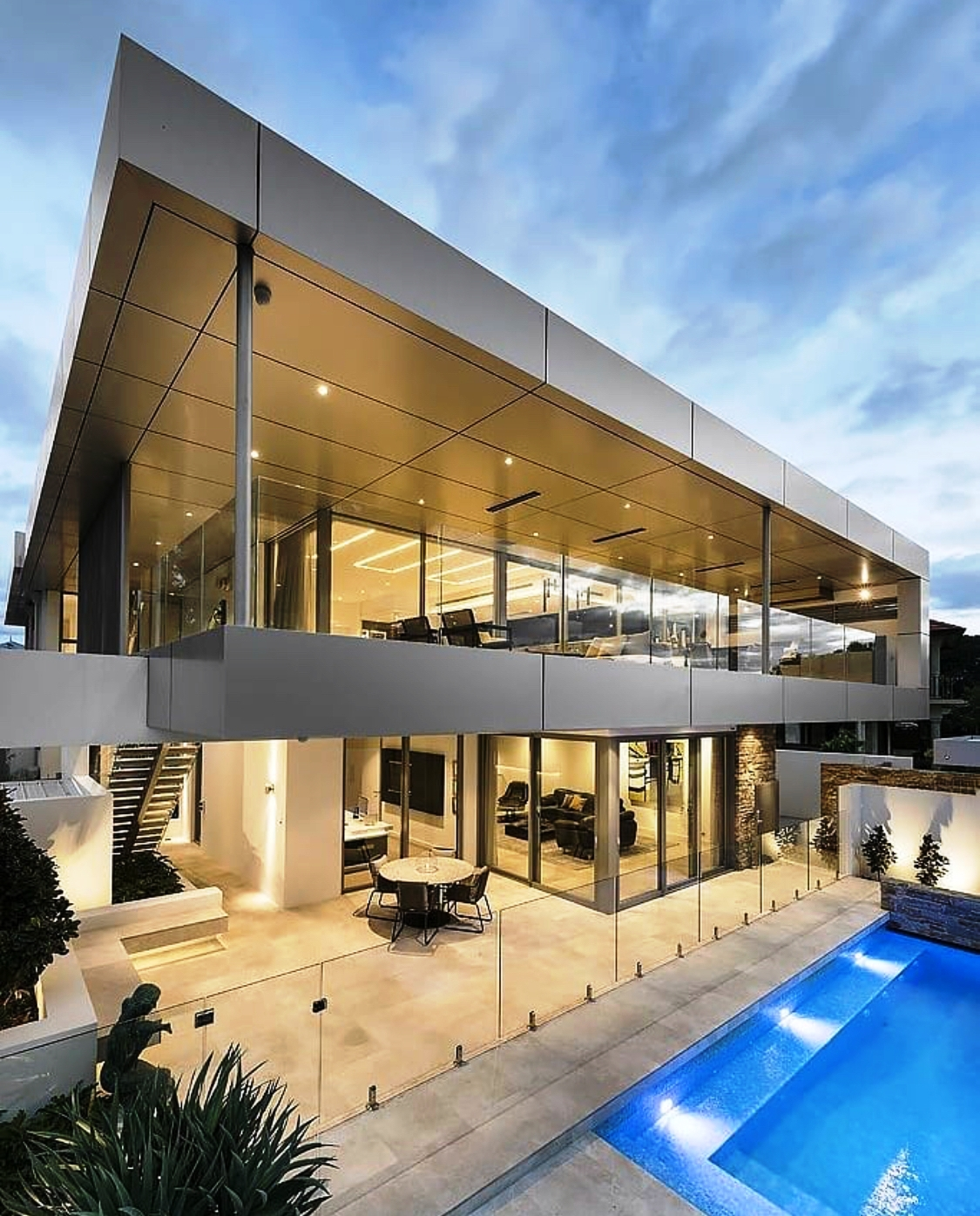 Modern, multi-story house with glass walls, outdoor patio, and swimming pool. Evening lighting.