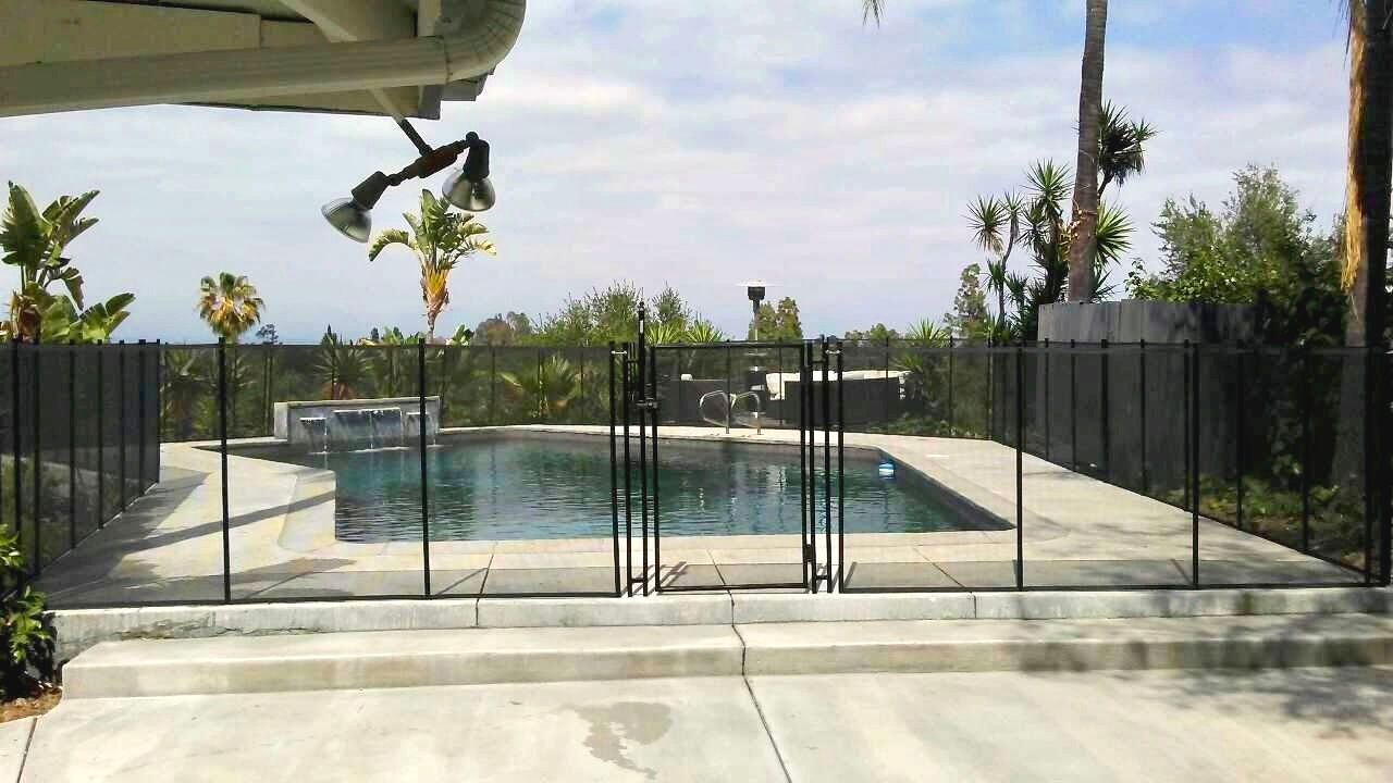 Pool enclosed by black mesh fence with gate, concrete patio, trees and sky background.