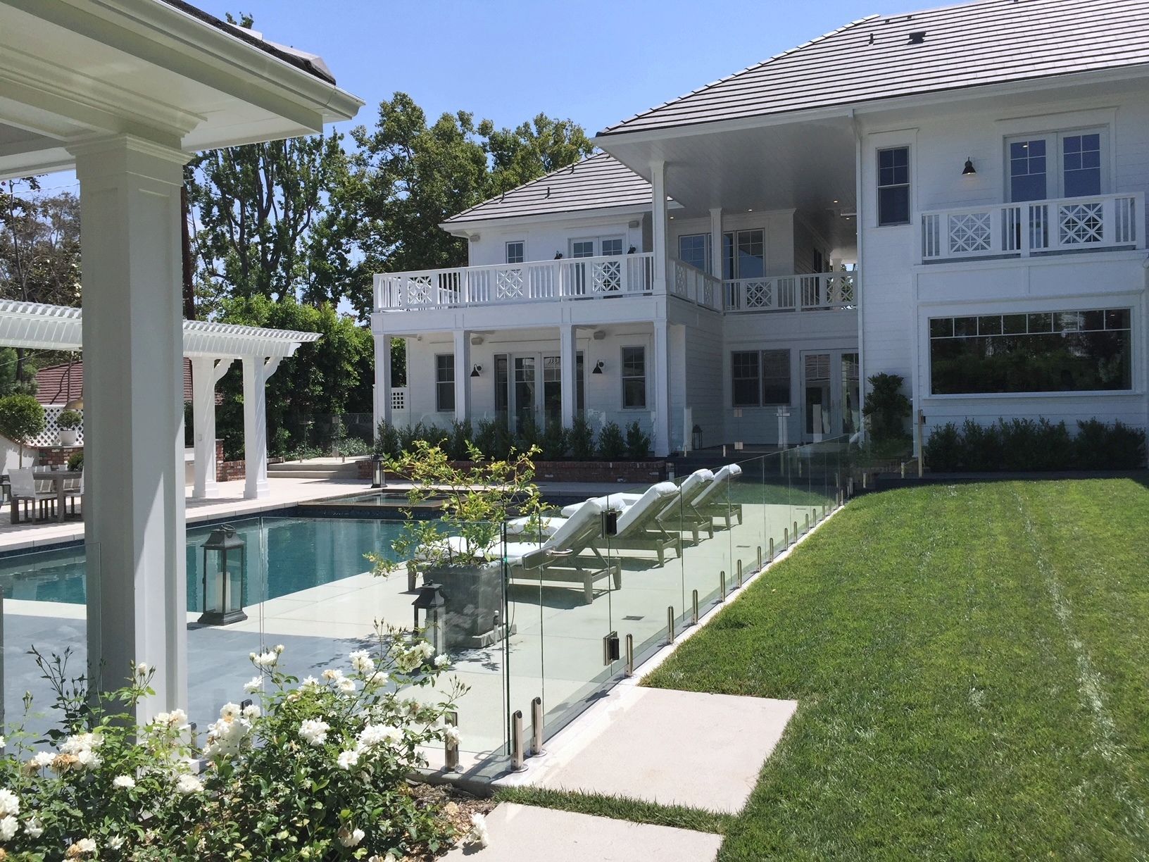 White house with pool, patio, and glass fence. Green lawn and sunny day.