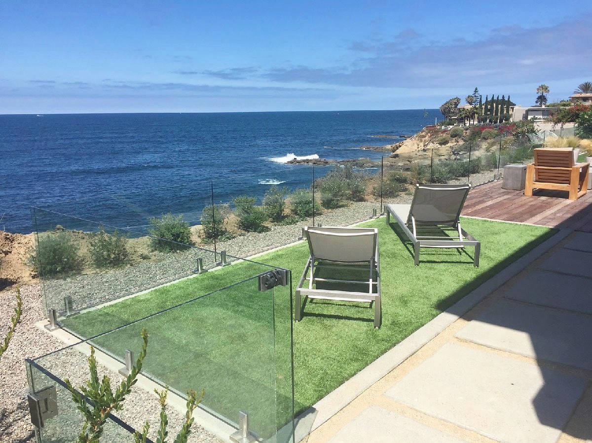 Oceanfront patio with two lounge chairs on artificial grass, glass railing, and blue ocean.