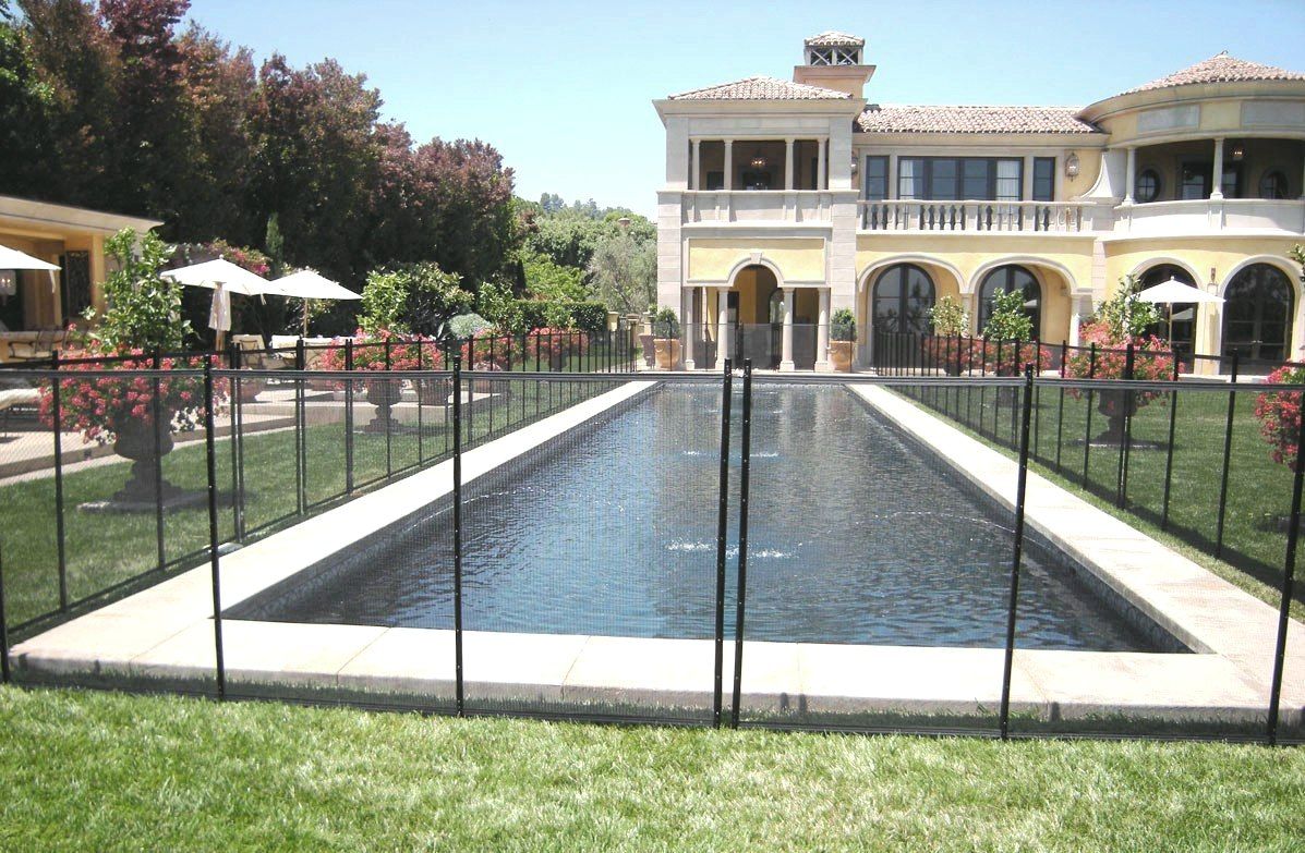 Pool with safety fence in front of a large, yellow mansion on a sunny day.