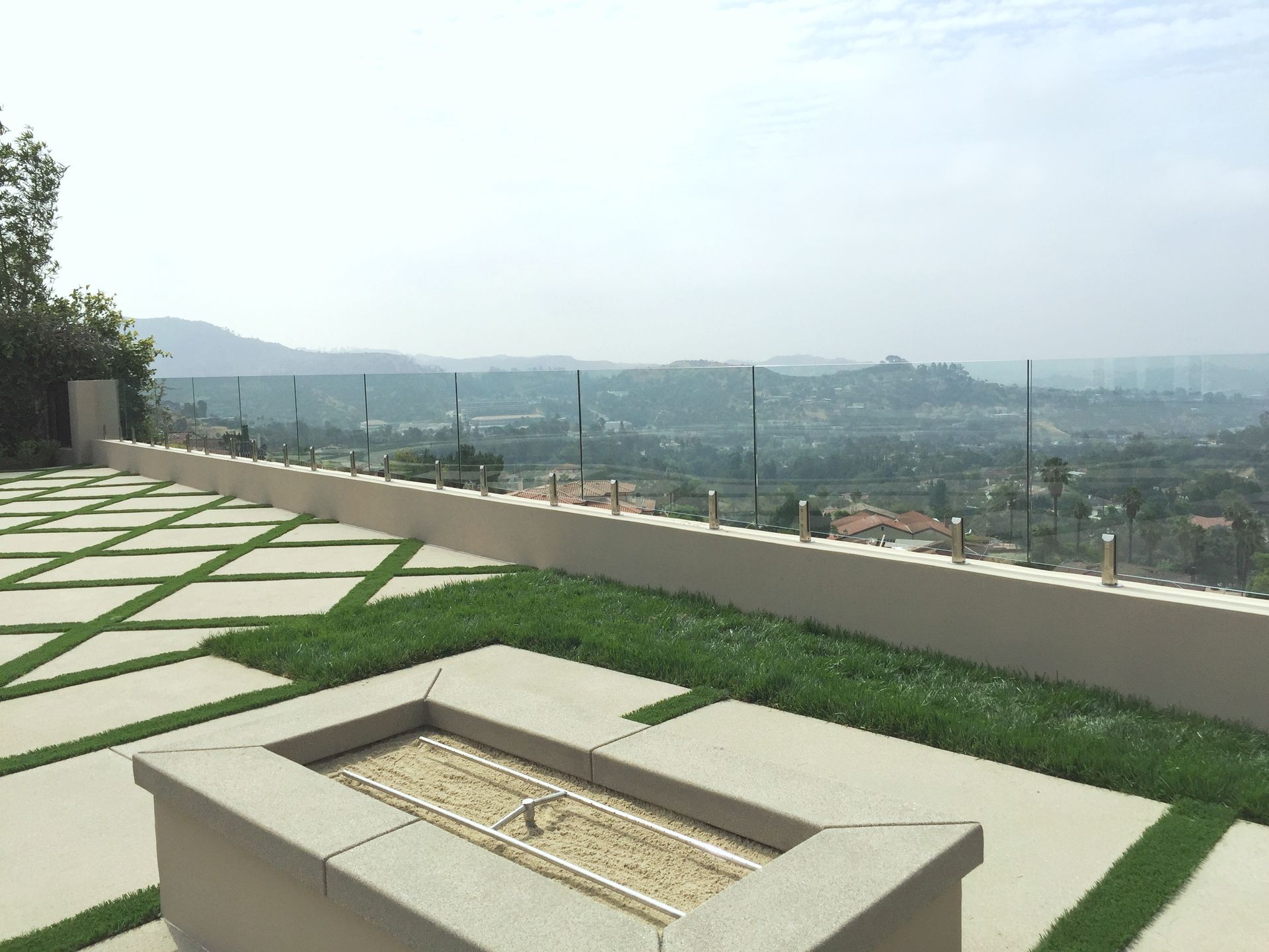Patio with concrete pavers, artificial turf, fire pit, and glass railing overlooking a valley.