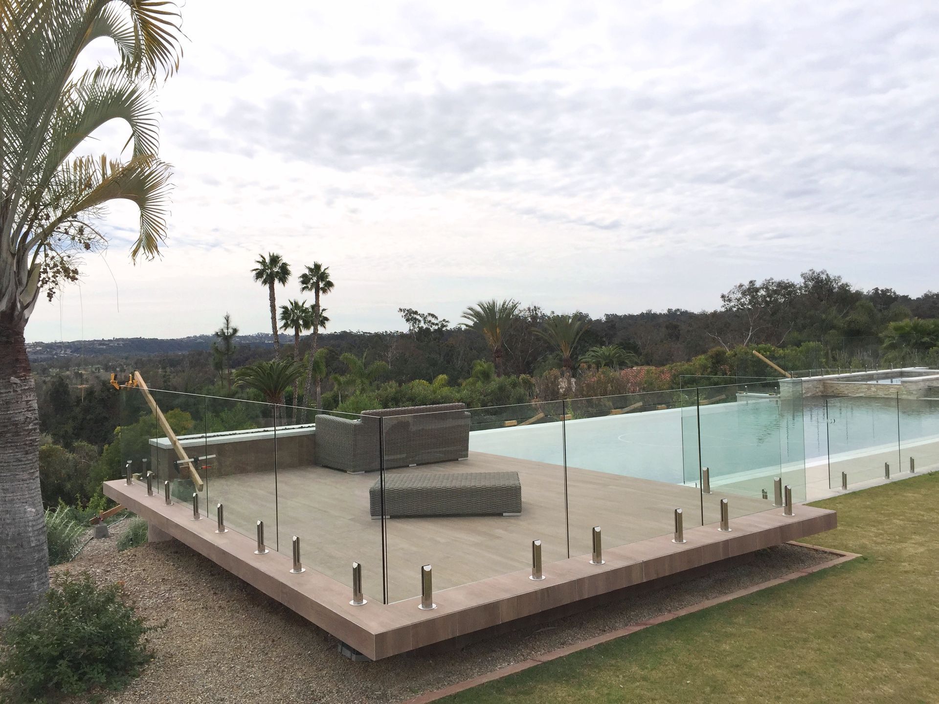 Deck with glass railing overlooking a pool and expansive landscape under cloudy skies.
