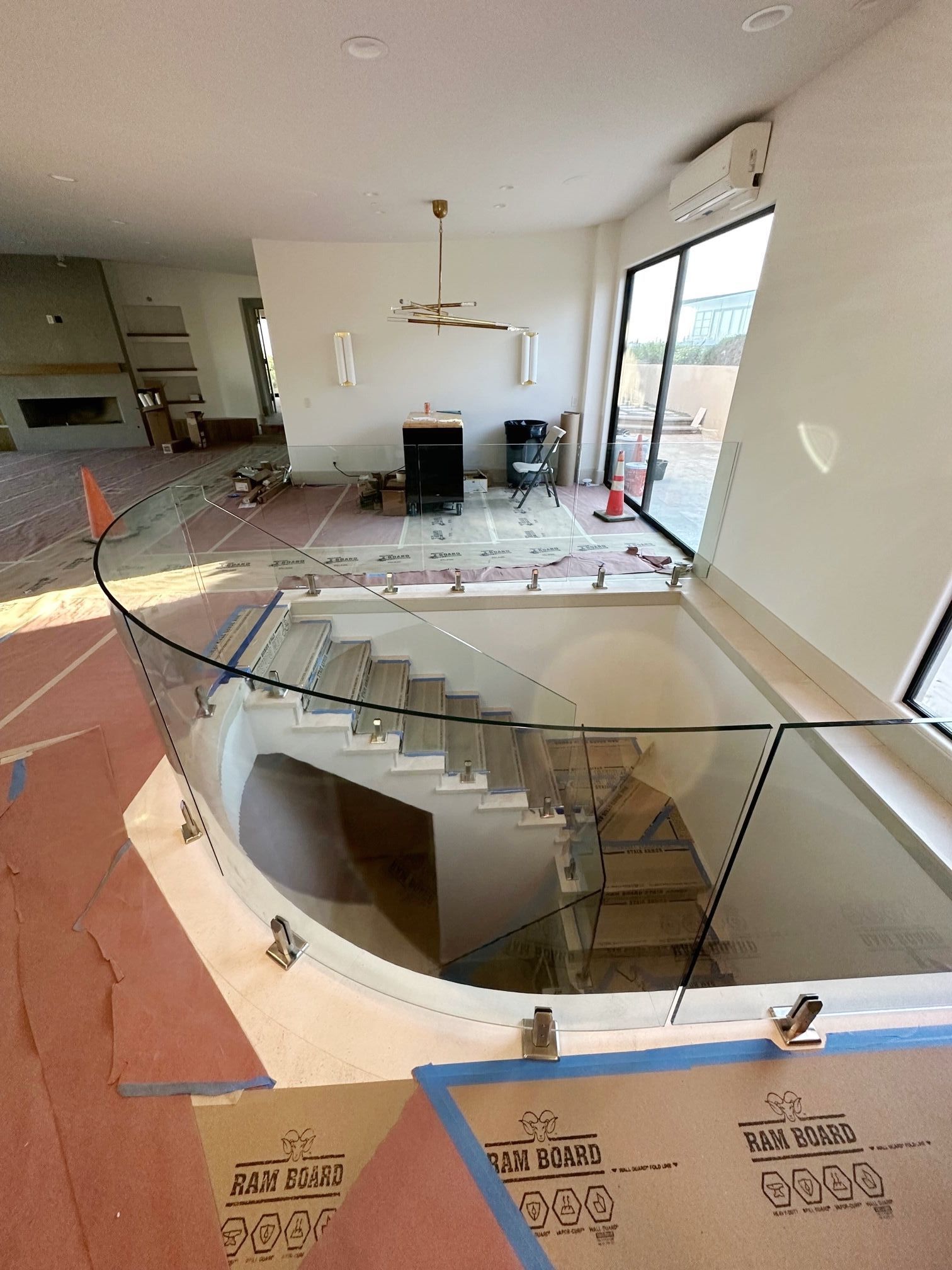 Interior view of a modern home with a curved staircase, glass railing, and large windows. Construction materials are visible.
