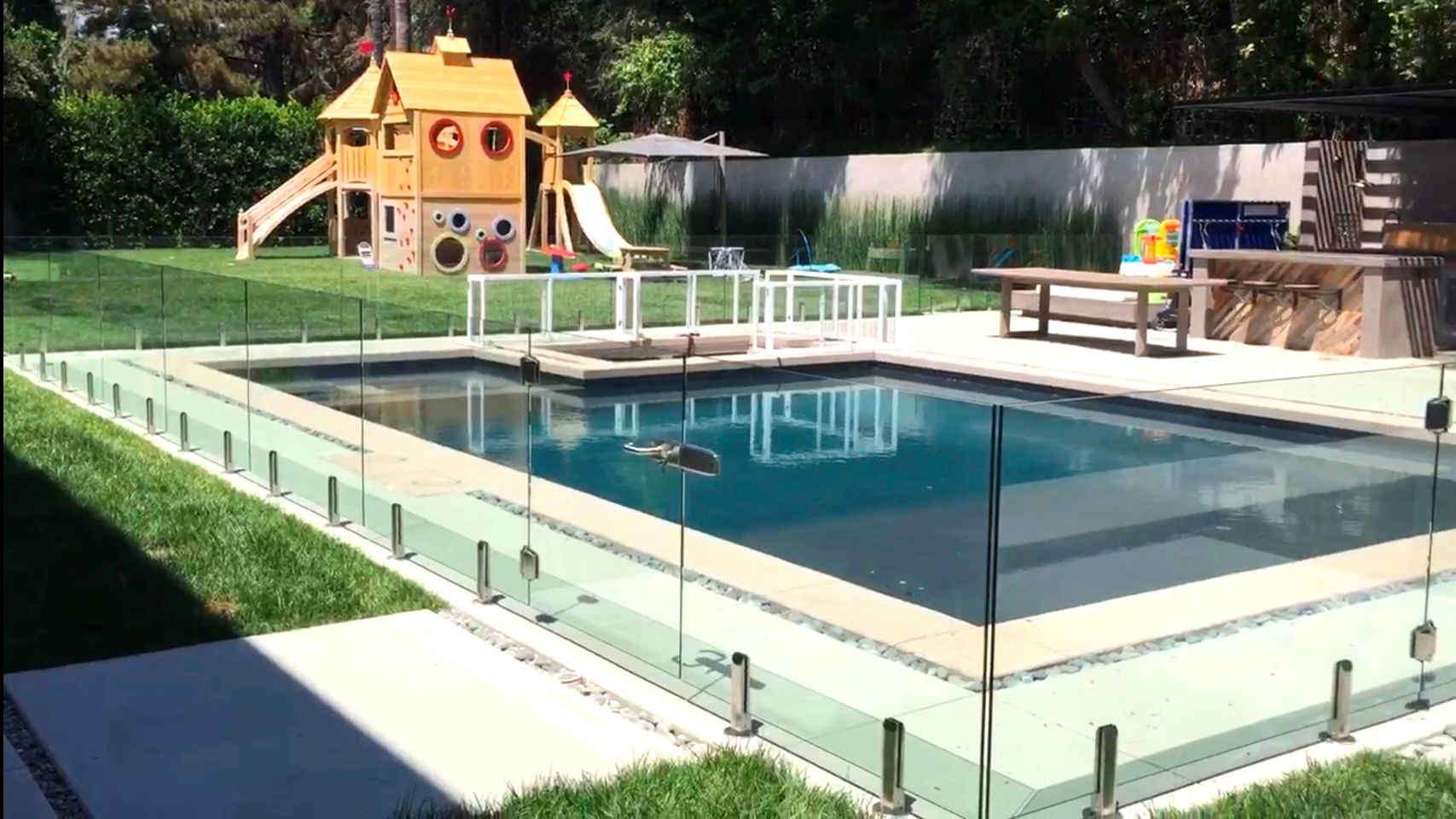 Rectangular pool with glass fence, small playhouse, and outdoor kitchen.