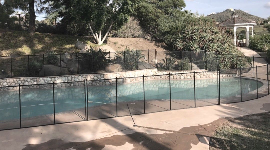 Black safety fence surrounds a curved swimming pool. Trees, shrubs, and a gazebo are in the background.