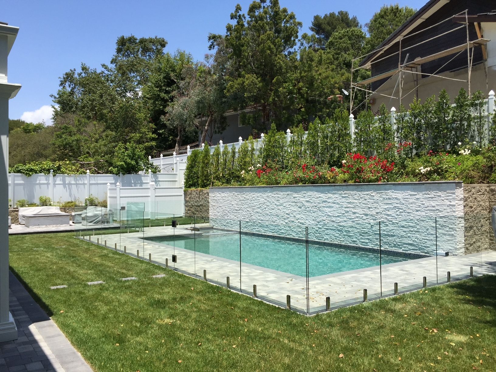 Rectangular swimming pool in a green lawn, surrounded by glass fencing. A stone wall with red flowers is behind the pool.