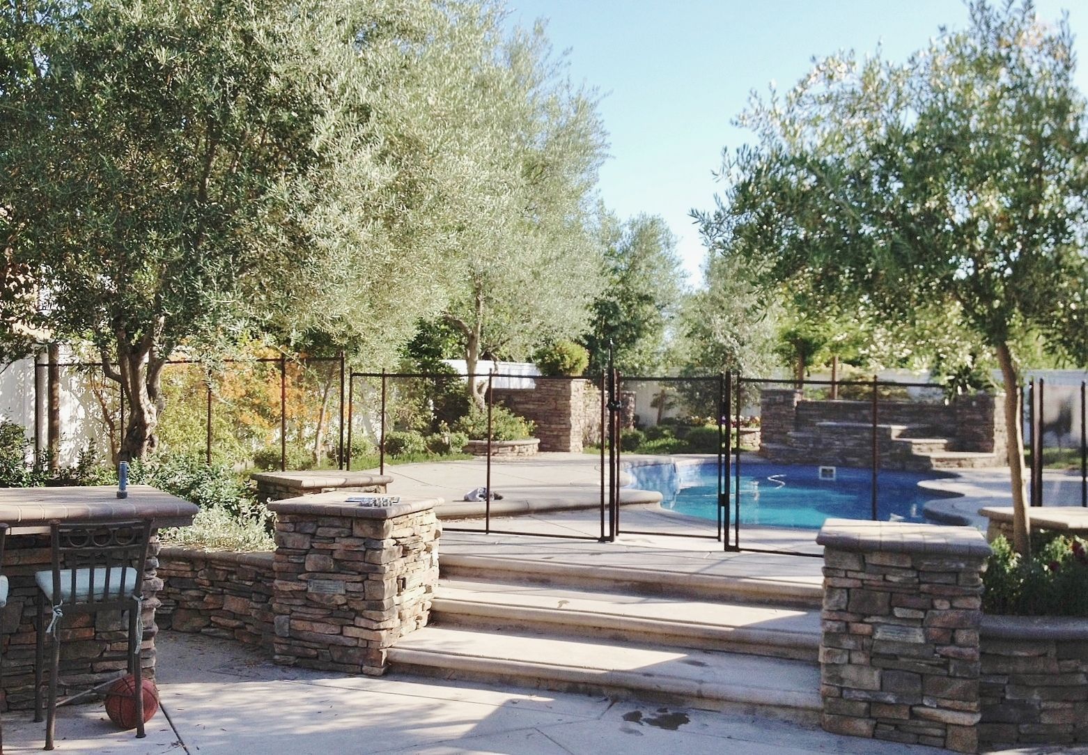 Stone-walled patio with steps leading to a pool, surrounded by trees and a black fence under a clear sky.