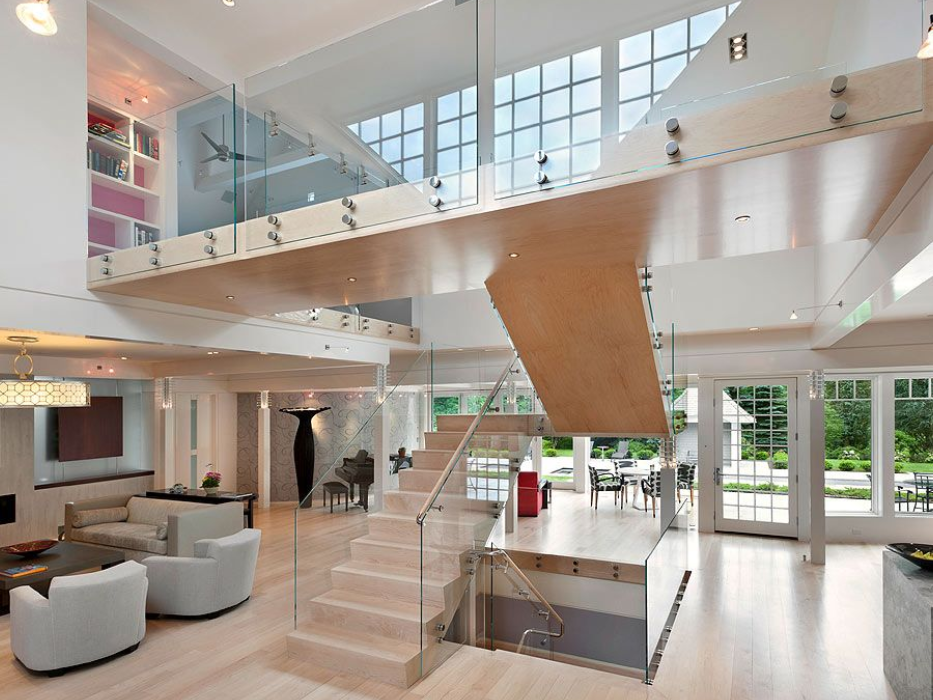 Modern home interior with mezzanine and glass staircase; light wood floors, white walls, and large windows.