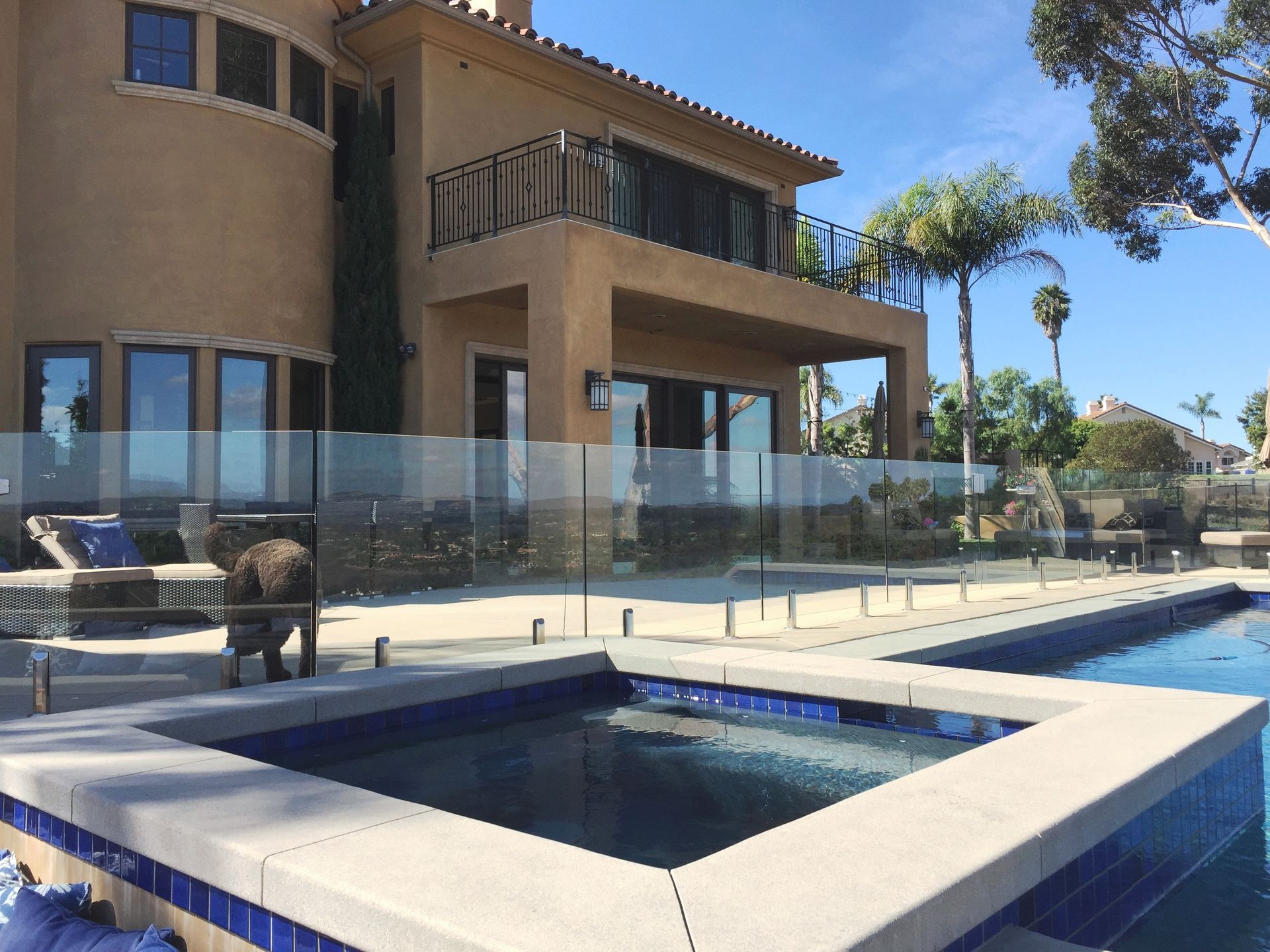 Backyard with pool, hot tub, and two-story house. Glass fence surrounds pool area. Sunny day.
