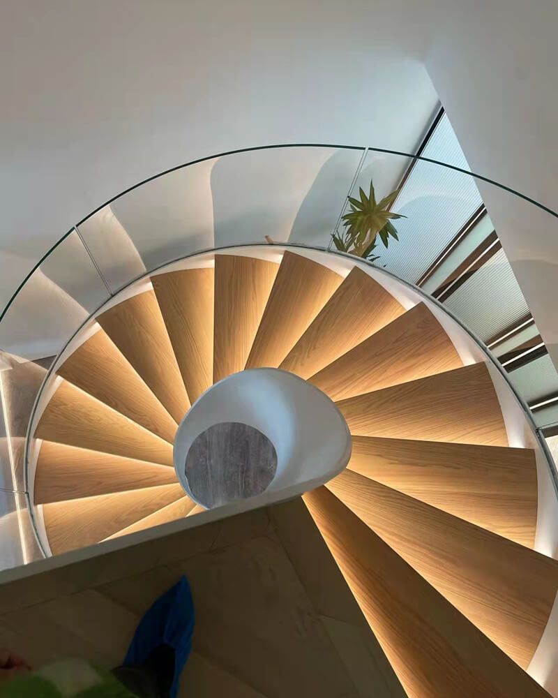 Spiral staircase with wooden steps lit from below, glass railing, and a person's foot in frame.