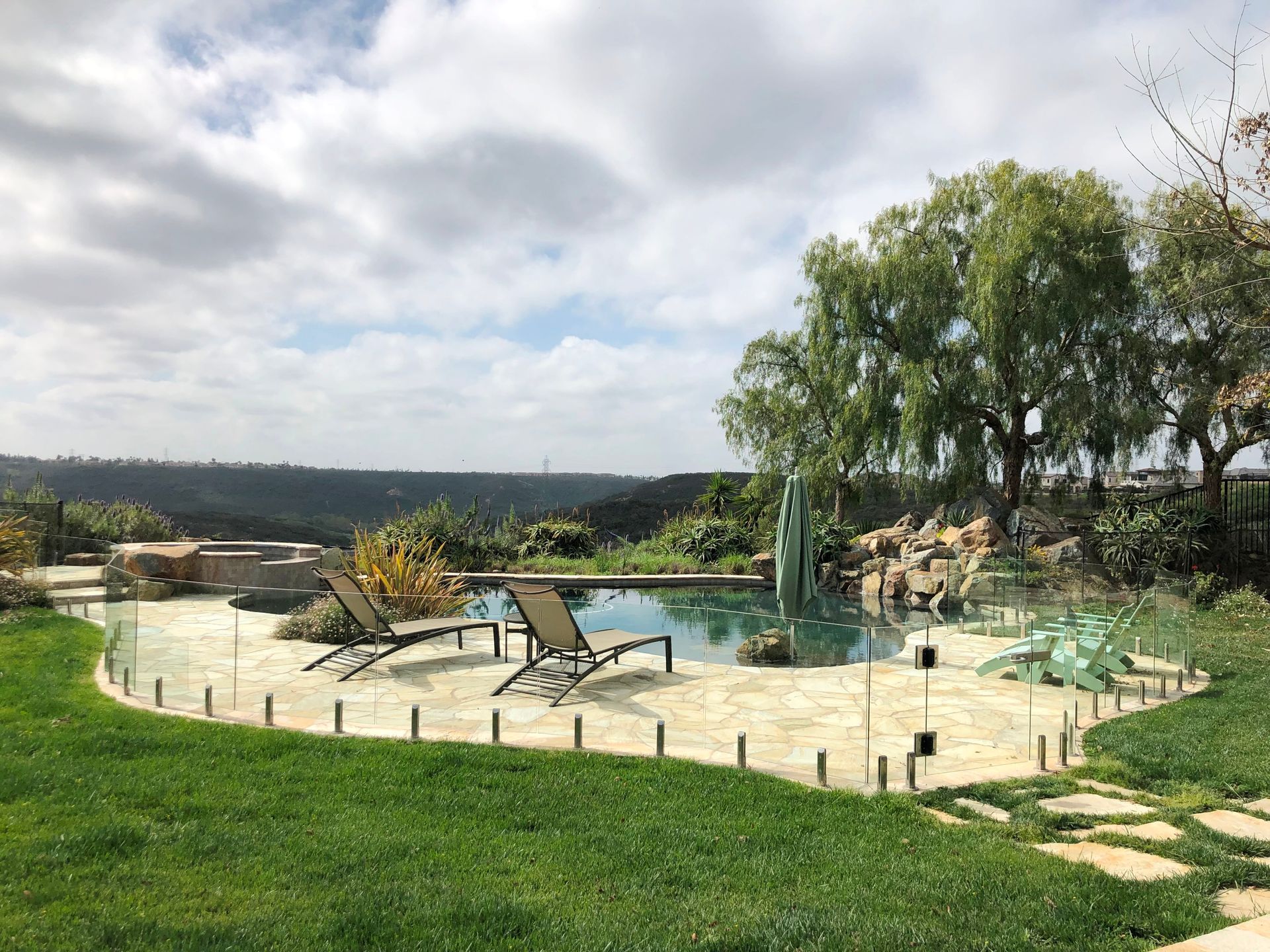 Pool with lounge chairs, stone patio, green lawn, and scenic view under a cloudy sky.