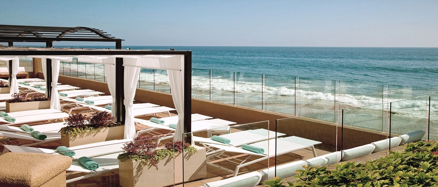 Lounge chairs on a beach with ocean view under a pergola, blue sky overhead.