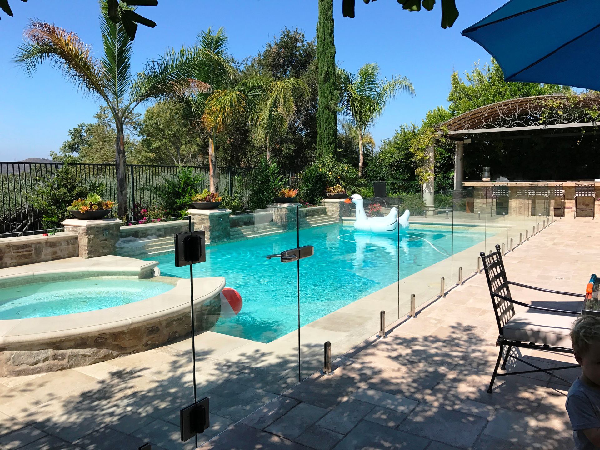 A sunny backyard pool with a jacuzzi, glass fence, and swan float.