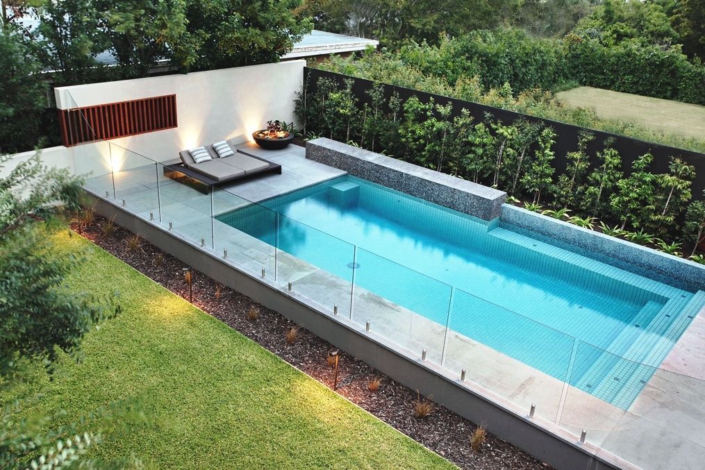 Modern backyard pool with lounge area, glass fencing, and lush green lawn.