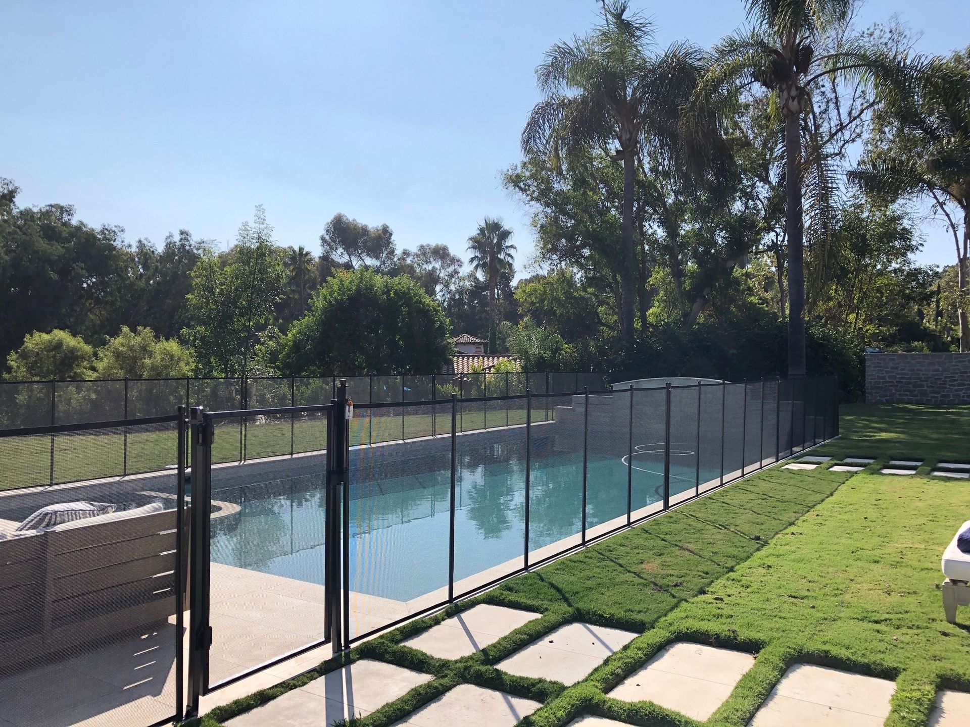 Swimming pool surrounded by a black mesh safety fence, green lawn, trees, and blue sky.