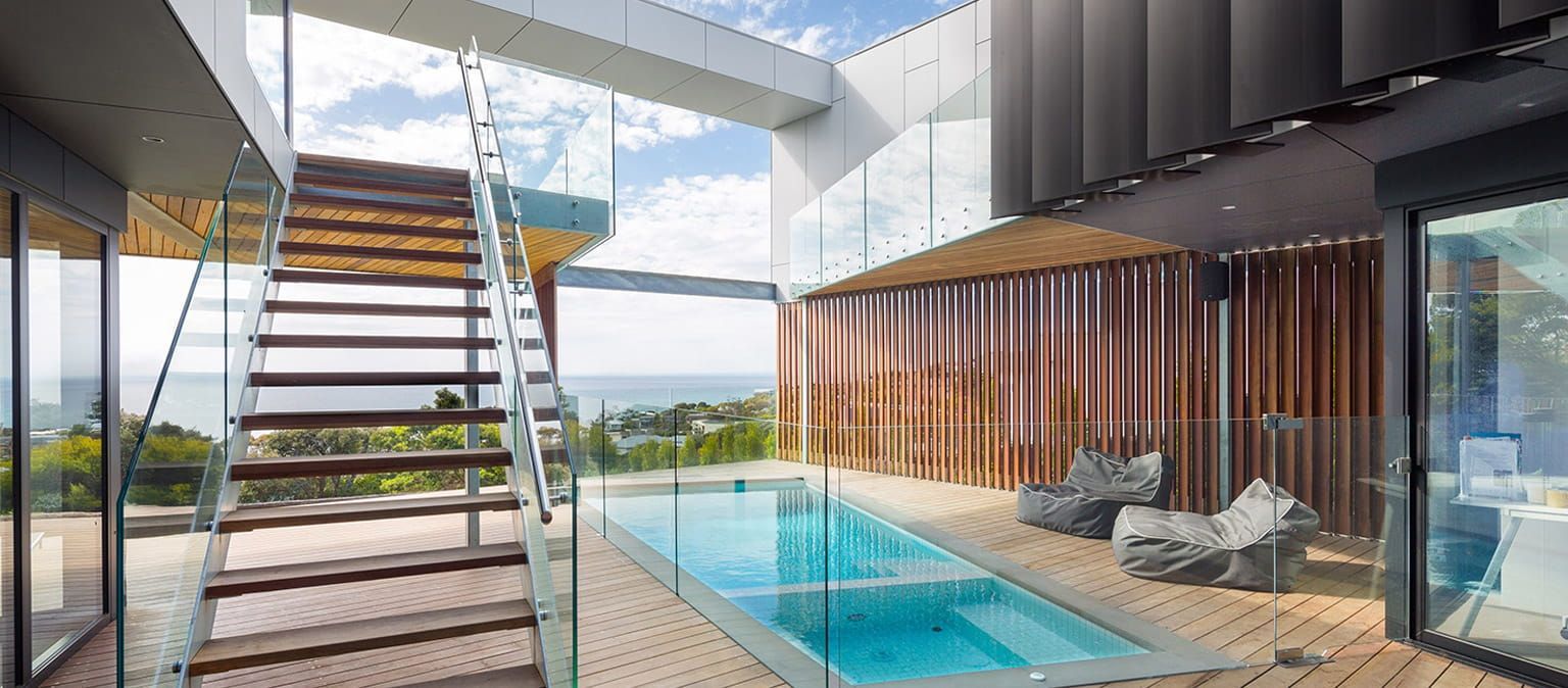 Modern home with a glass-enclosed pool, wooden deck, and staircase overlooking the ocean.