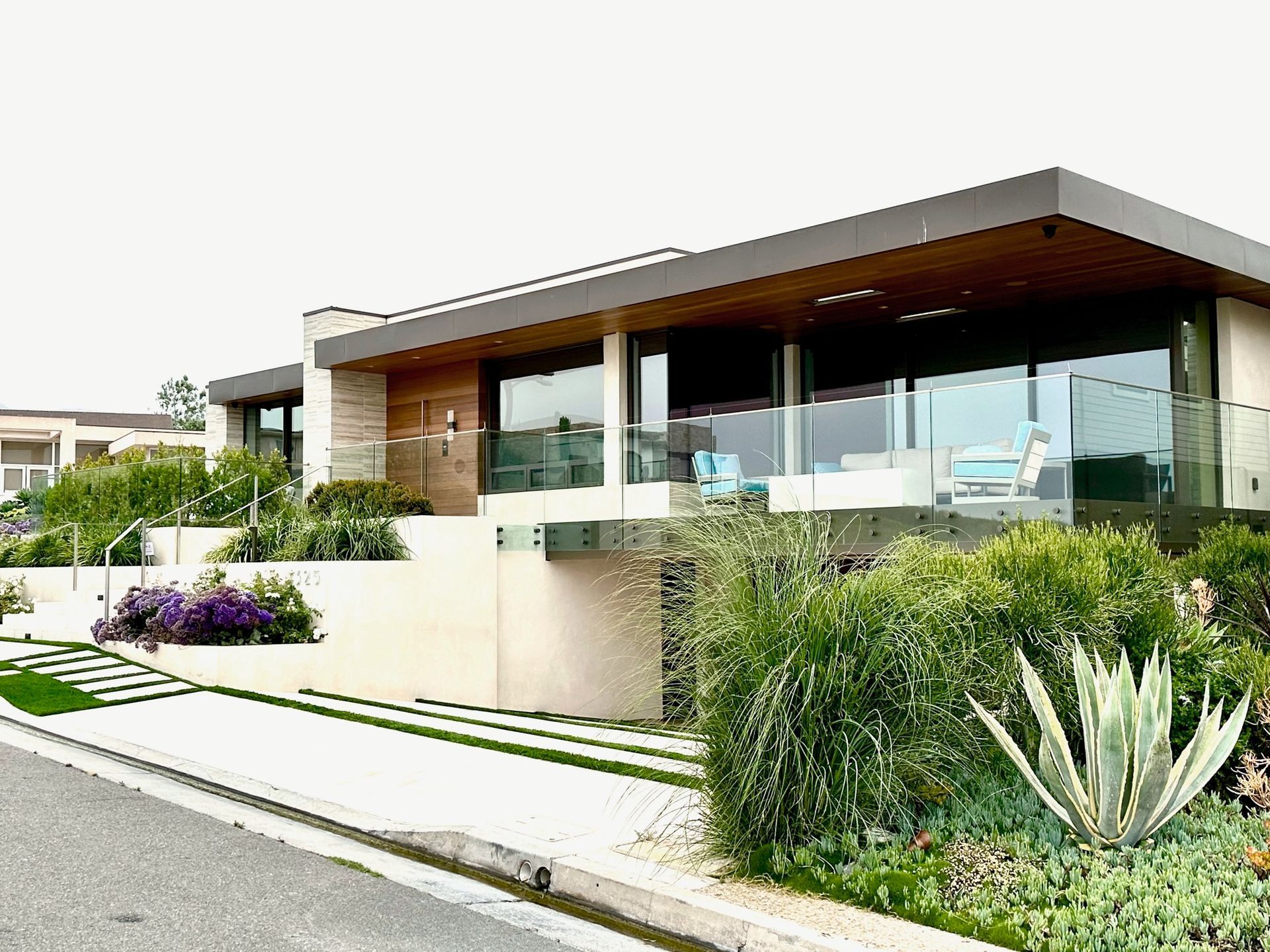 Modern house with large windows, wooden accents, and a glass-railed balcony; landscaping and driveway.