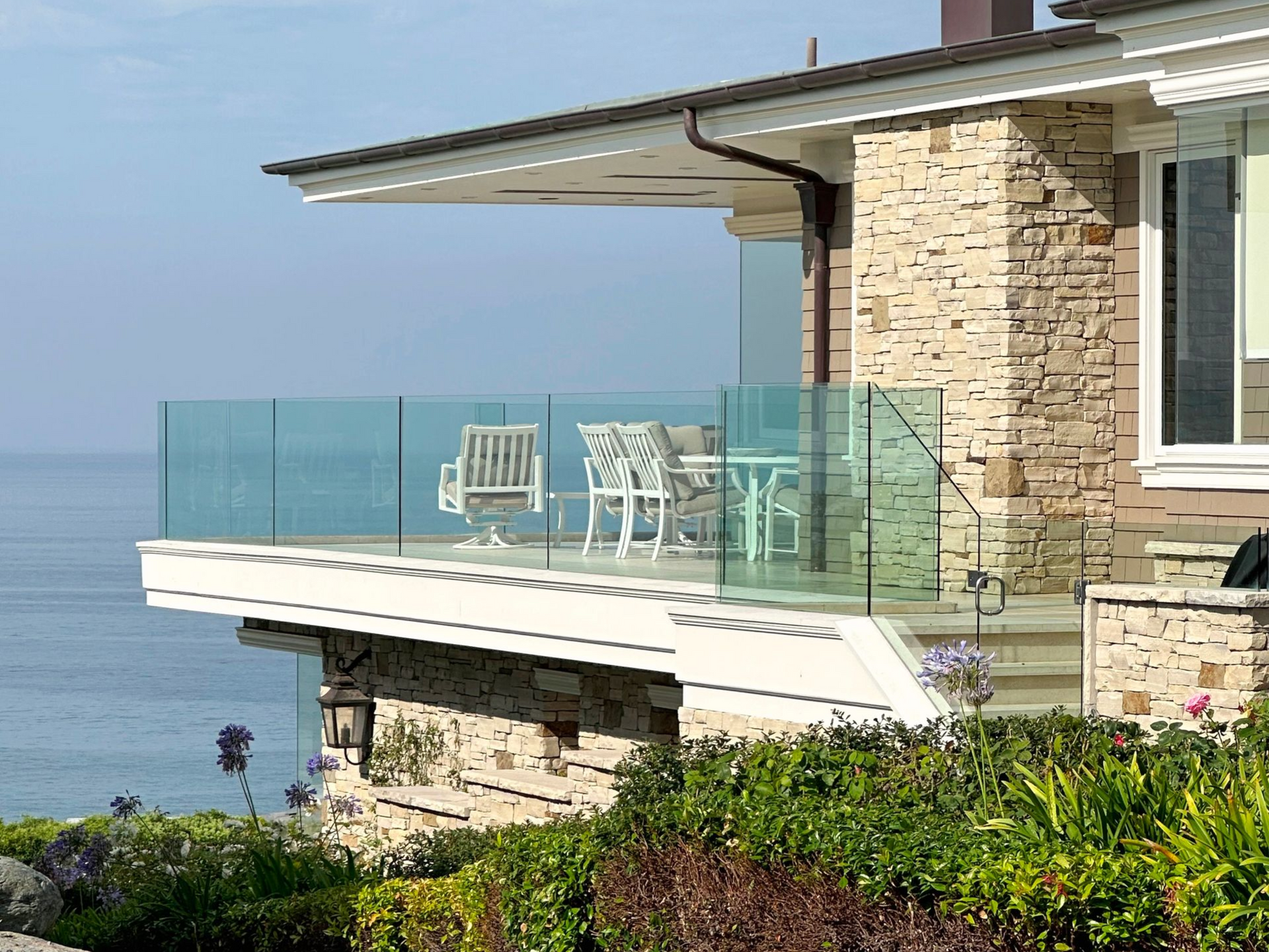 Glass-railed balcony with white furniture, overlooking the ocean. The house is stone with a brown roof.