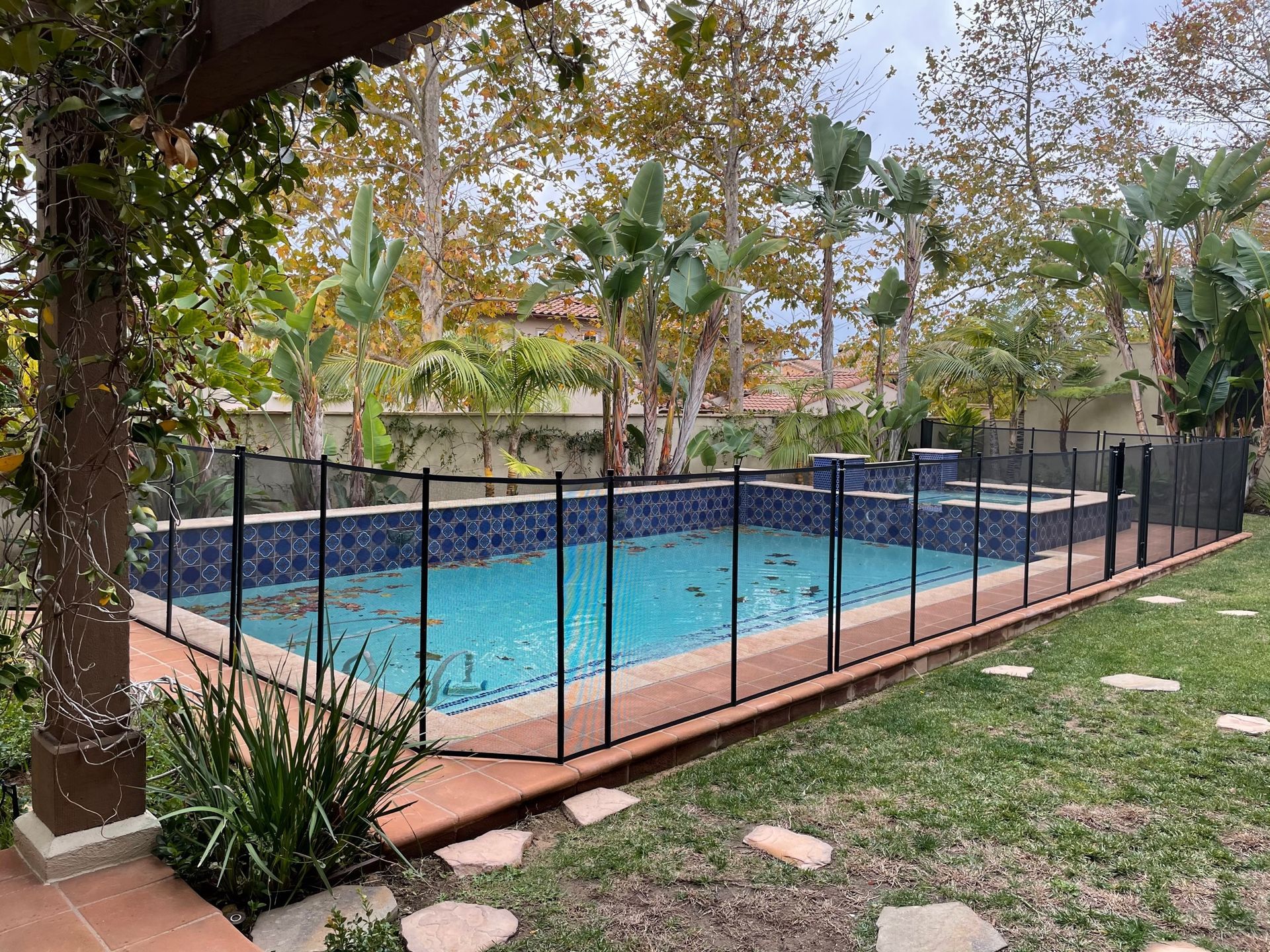 A swimming pool enclosed by a black safety fence in a backyard with trees.