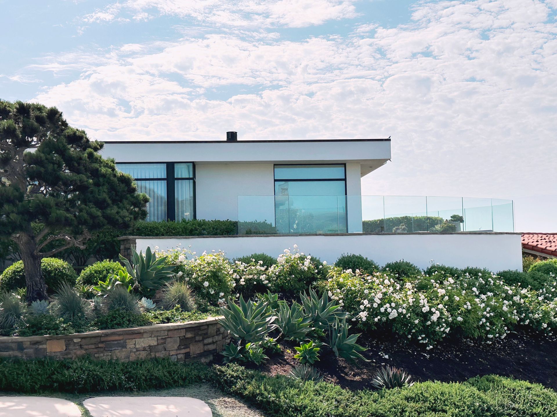 Modern white house with large windows and glass balcony overlooking lush landscaping.
