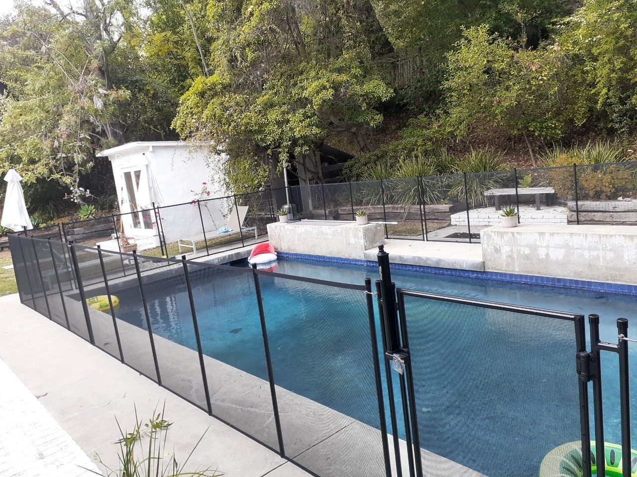 Pool area with a safety fence, small white building, and trees on a hillside.