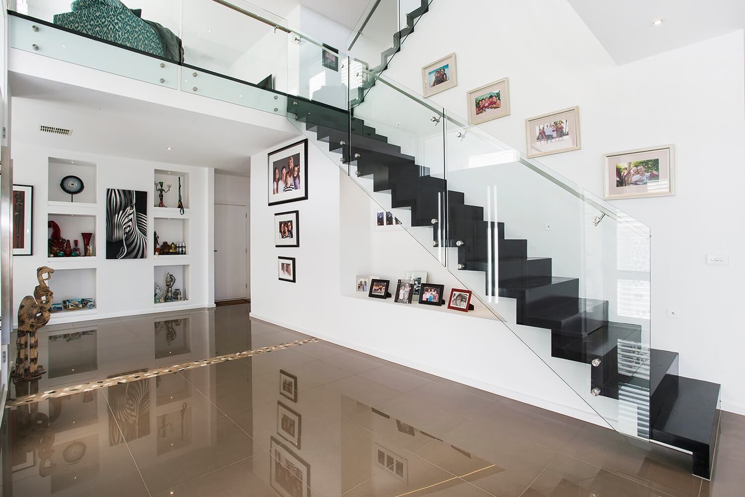 Modern home interior with a staircase, glass railings, and artwork on white walls. Brown shiny floor.