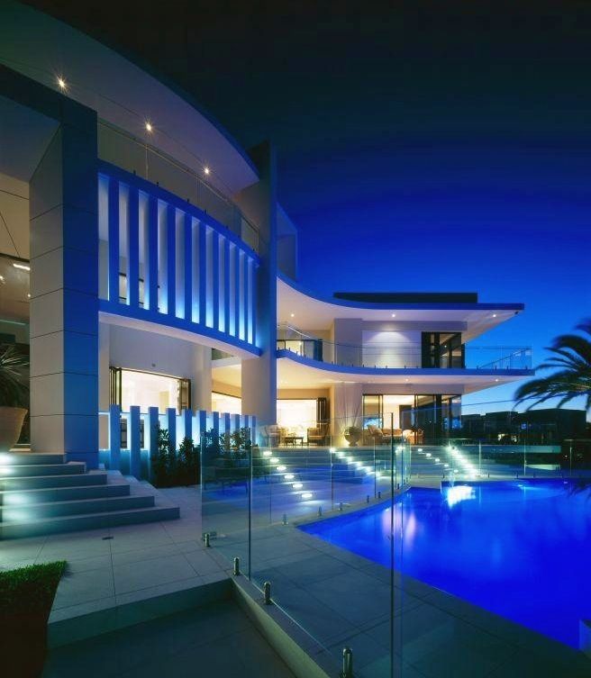 Modern luxury home with a blue-lit pool, stairs, and curved balconies at night.
