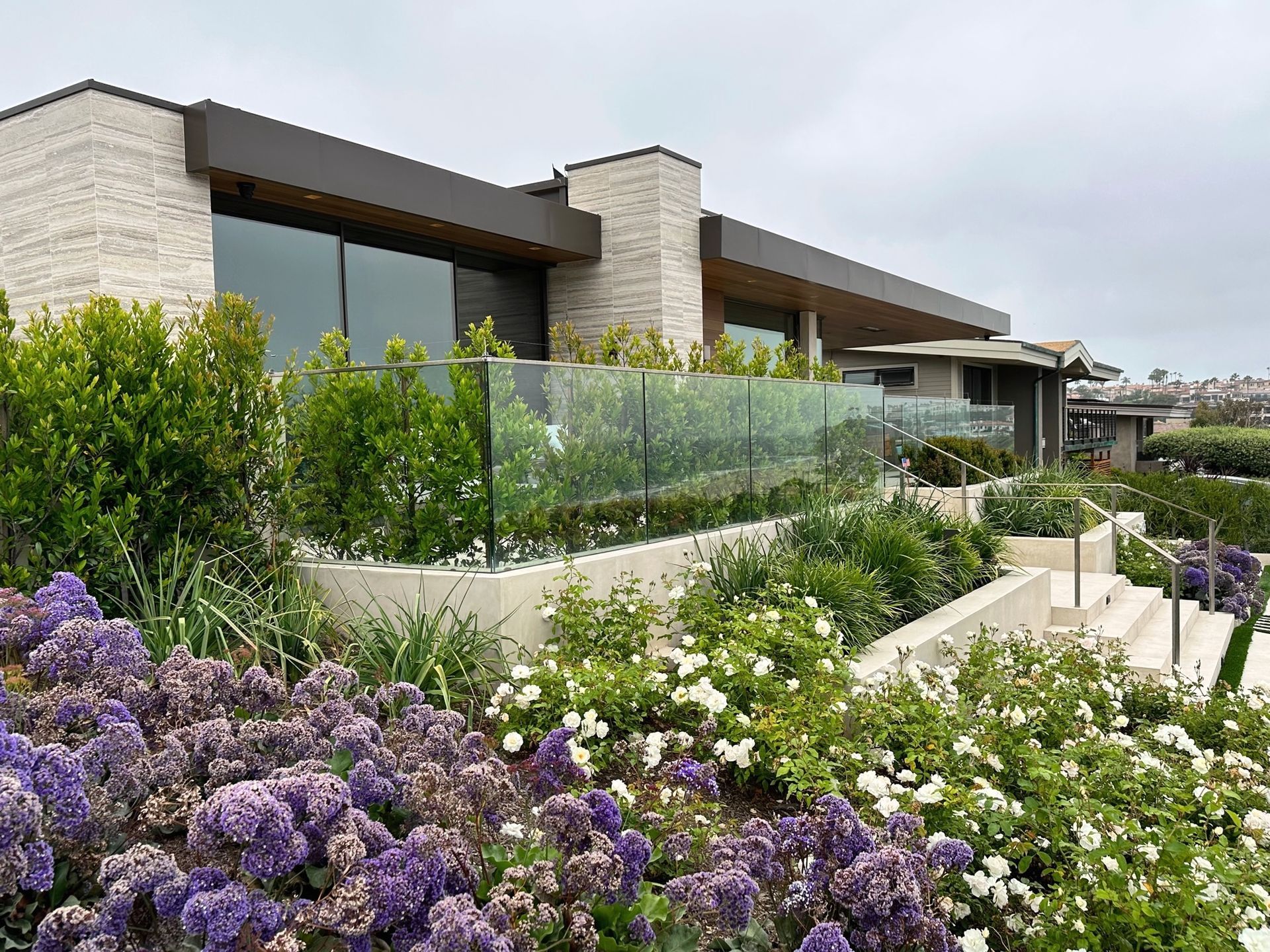 Modern house with glass railing, tiered garden with purple and white flowers, cloudy day.