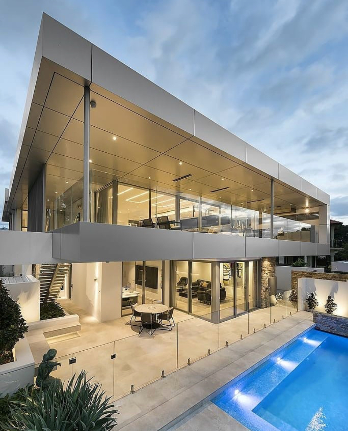 Modern two-story house with glass walls, outdoor patio, and a pool. Exterior is white and gray, with an overcast sky.