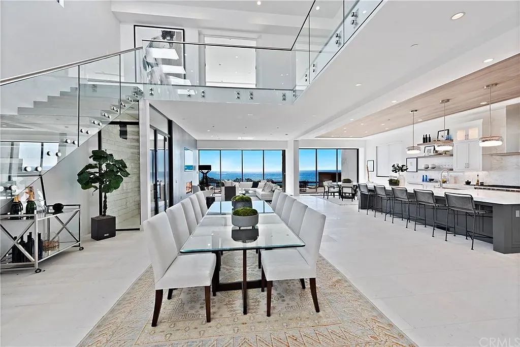 Spacious, modern dining area with a long glass table, chairs, and ocean views. A staircase and open kitchen are also visible.