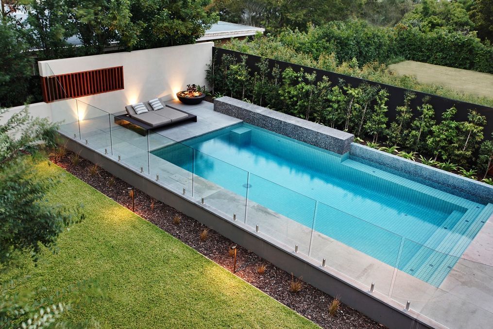 Modern rectangular swimming pool with glass barrier, chaise lounge, and fire pit in a manicured backyard.