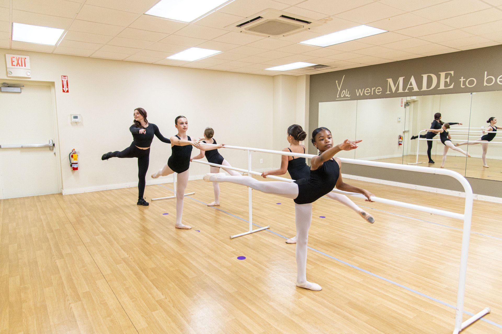 Dancers in leotards at barre in dance studio. Wooden floor, mirrors, white barre and wall.