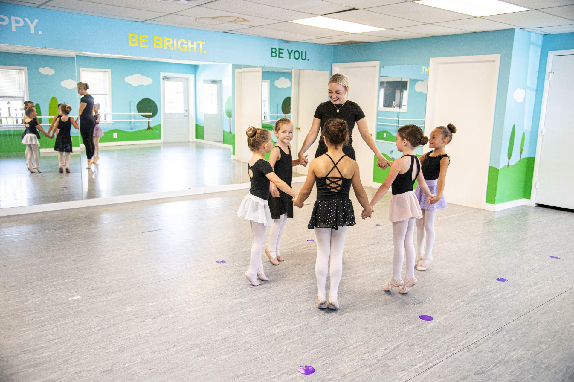 Children in ballet attire circle holding hands with a teacher in a dance studio.