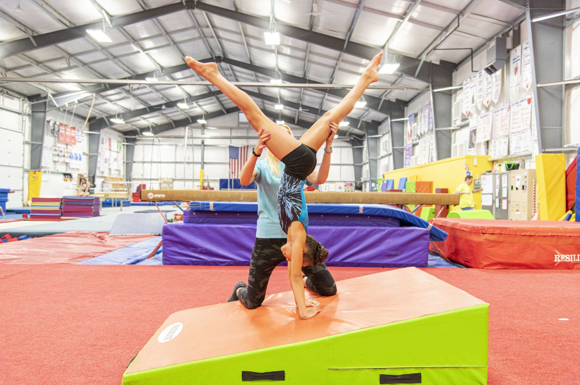 Two gymnasts: one upside down on a block, supporting another in a split in a gym.