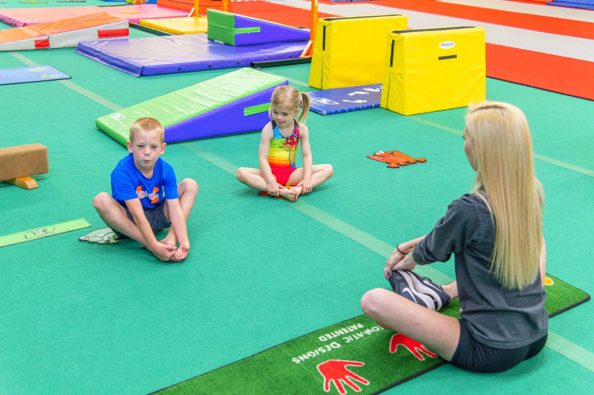 Boy and girl sit on floor, instructor facing them. Gymnastics gym setting, green floor, mats, colorful equipment.