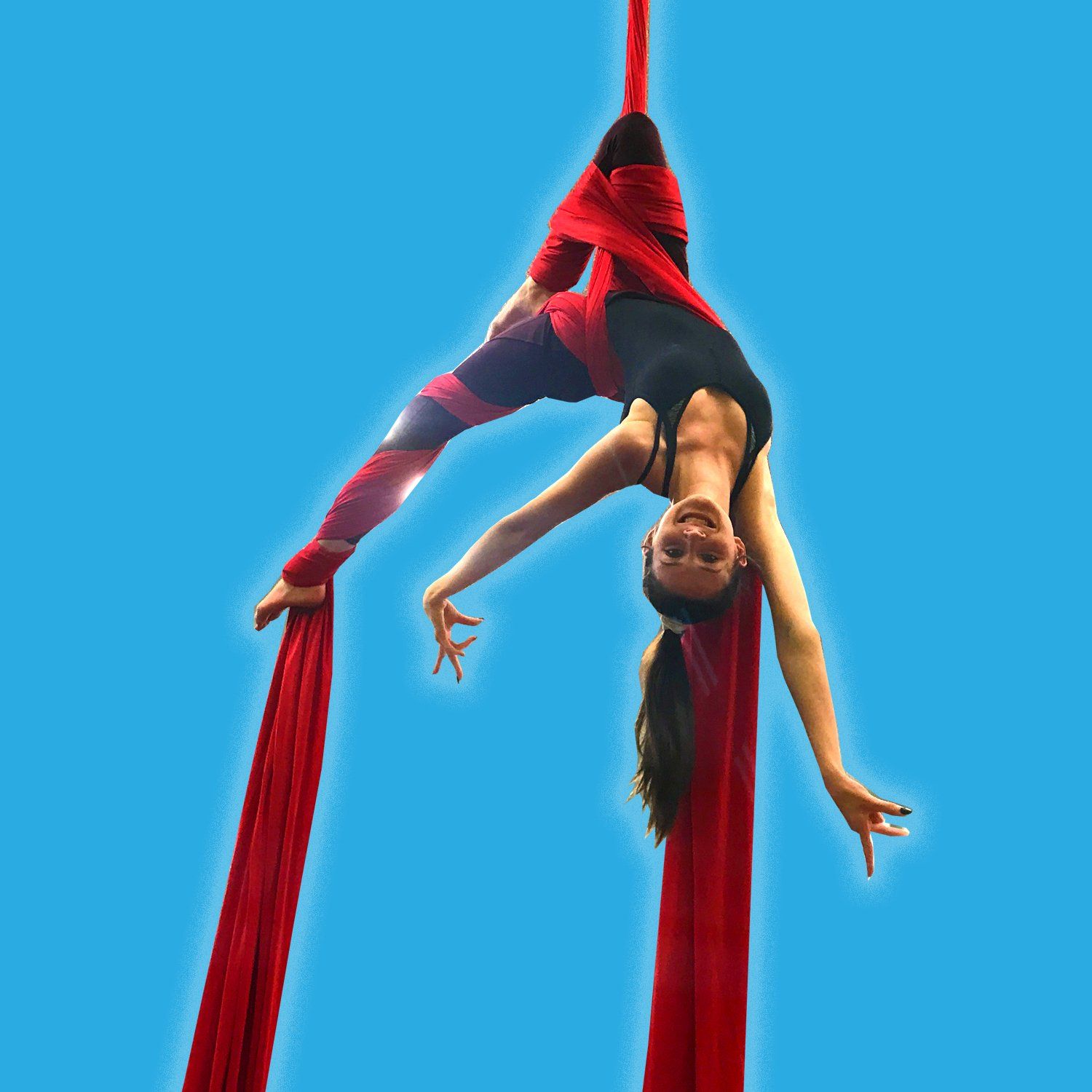 Woman upside down on red aerial silk, smiling against a blue sky.