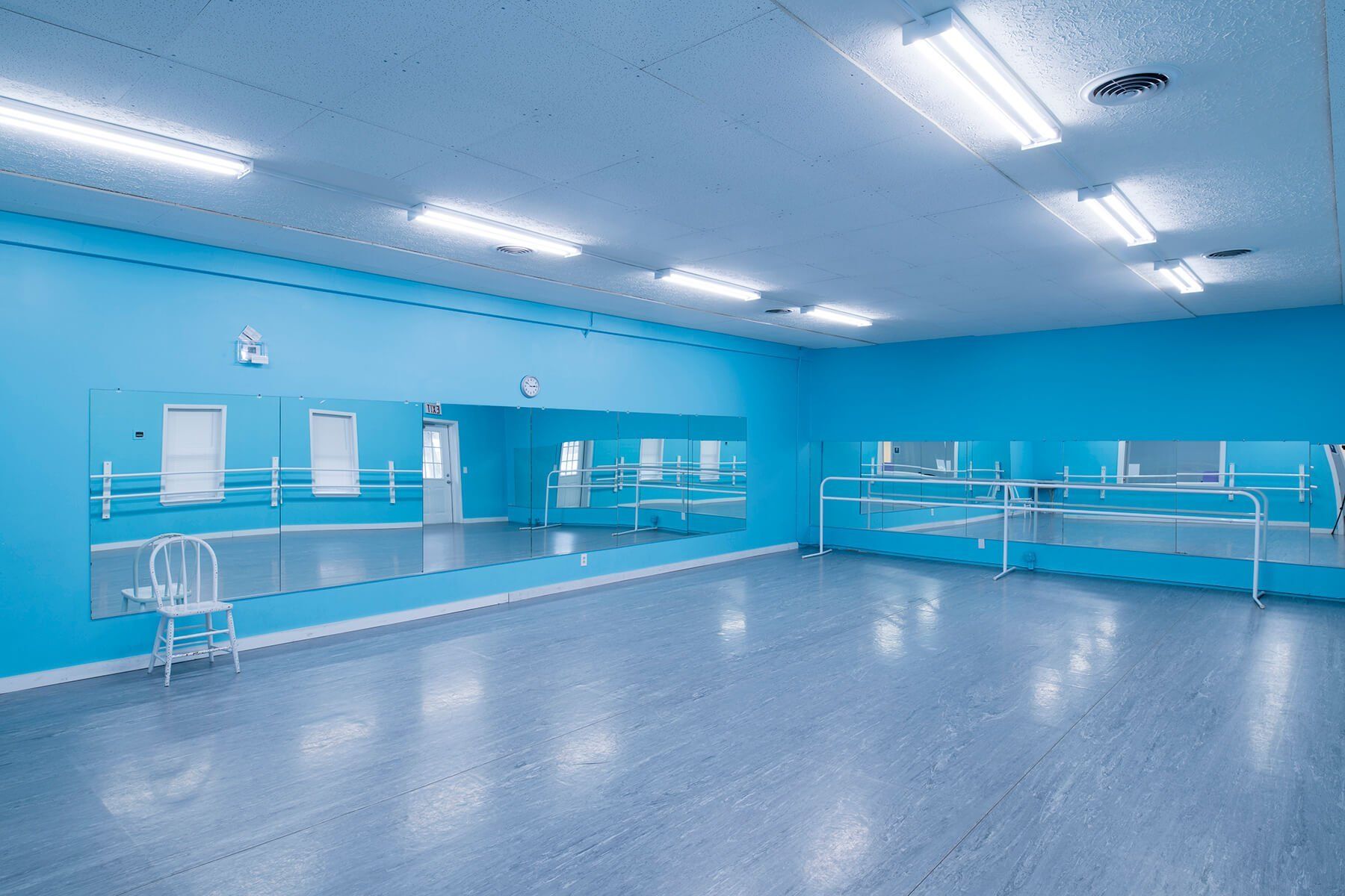 Blue-walled dance studio with mirrored wall, barre, and white chair. Fluorescent lighting illuminates the room.