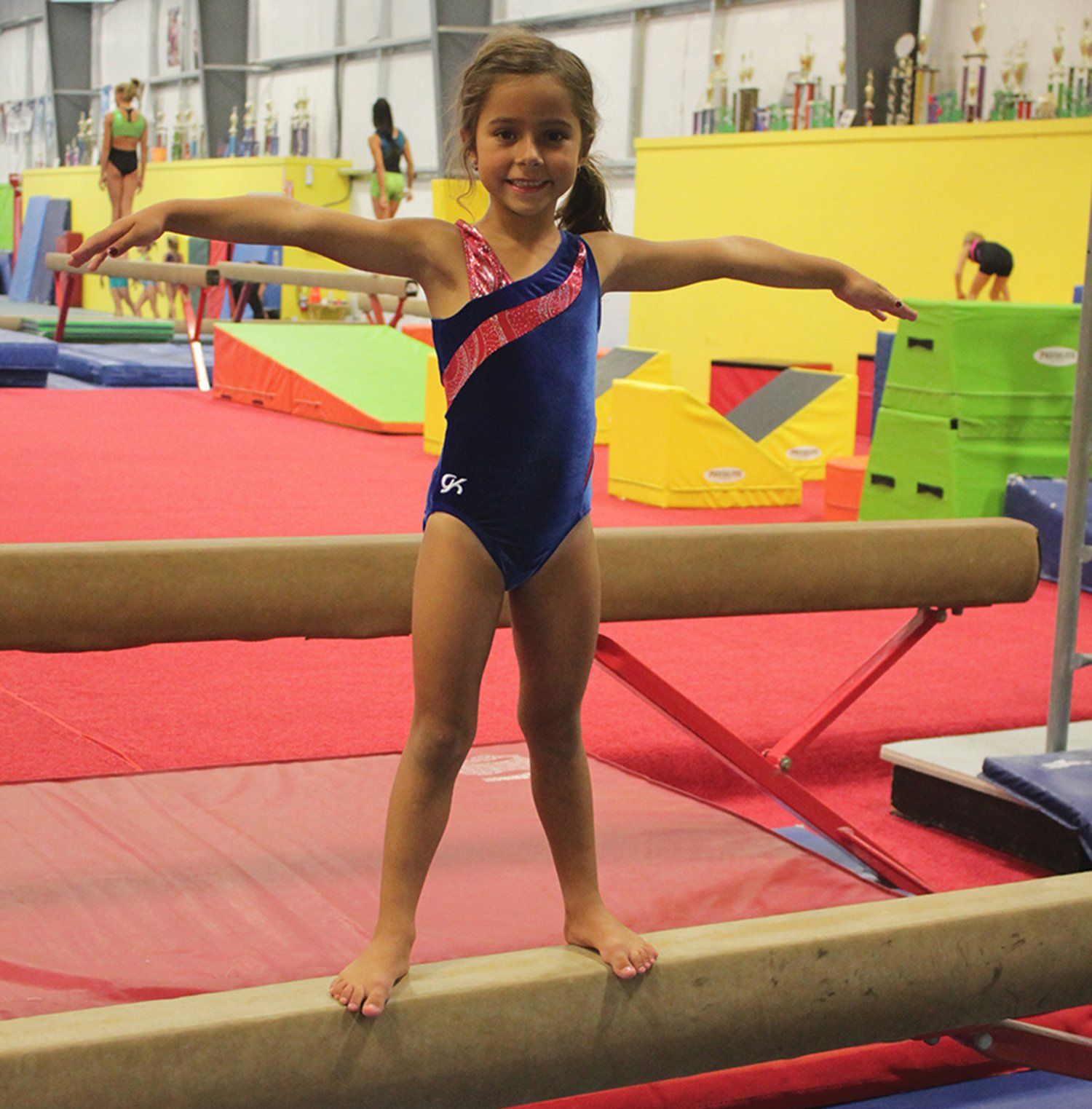 Girl in a blue leotard balancing on a beam with arms out in a gymnastics gym.