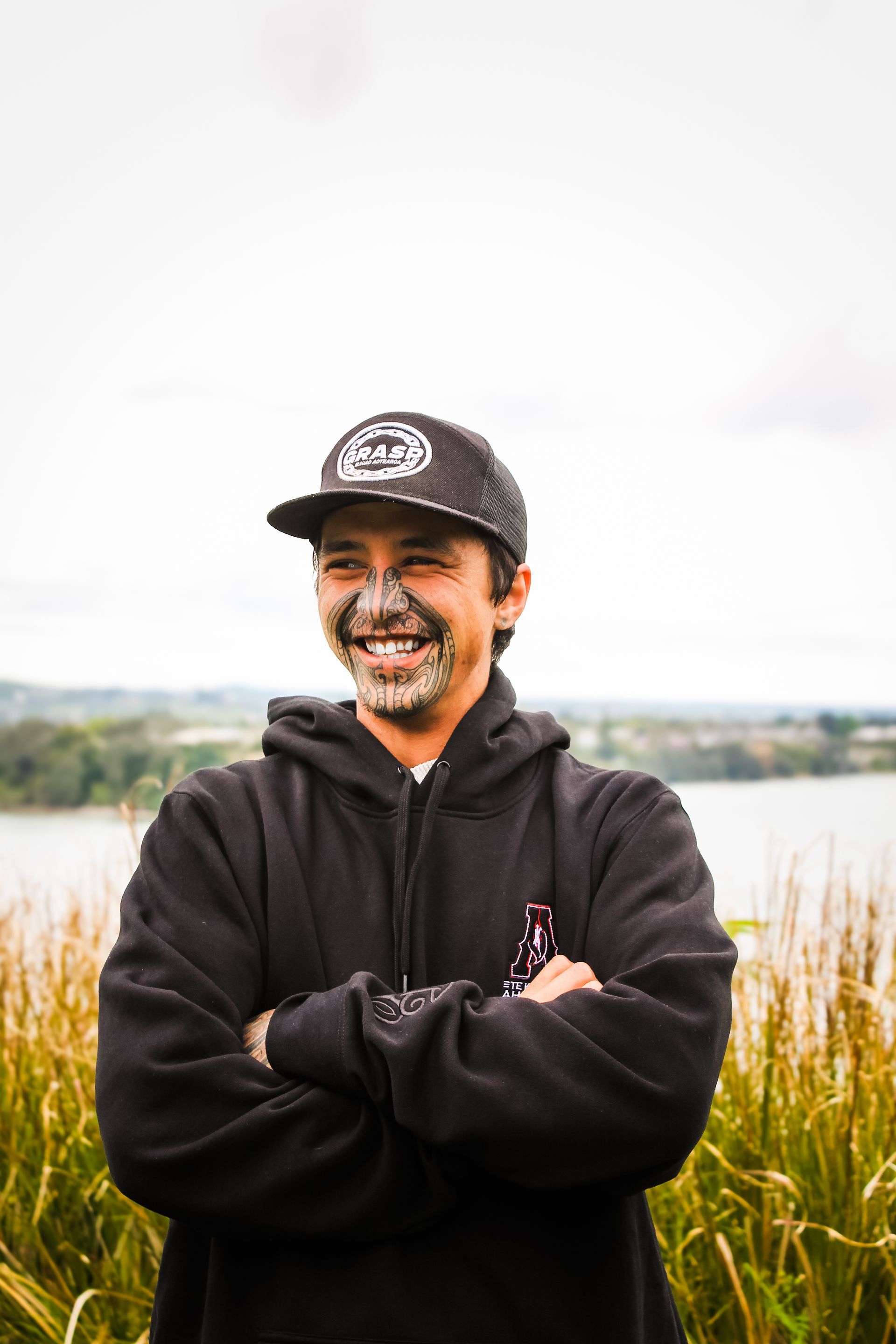 A man wearing a hat and a hoodie is standing in a field with his arms crossed.