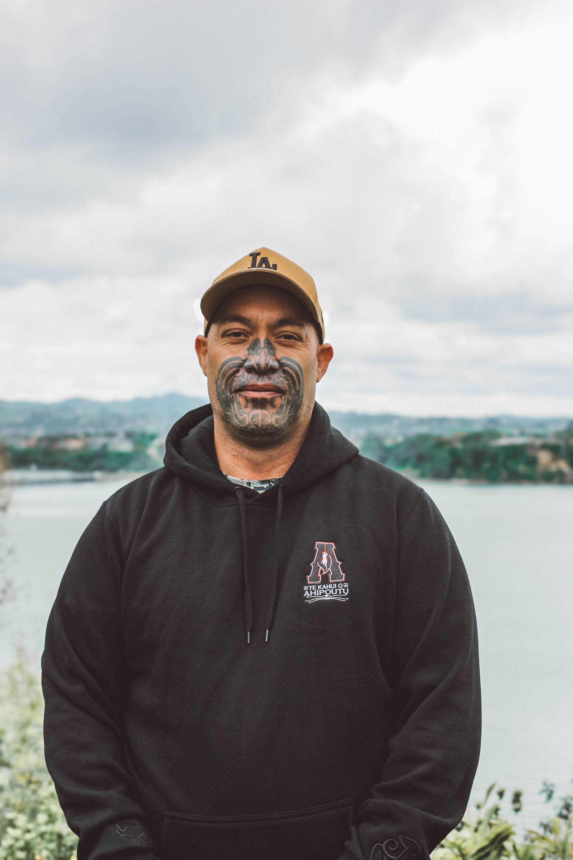 A man with a tattoo on his face is standing in front of a body of water.
