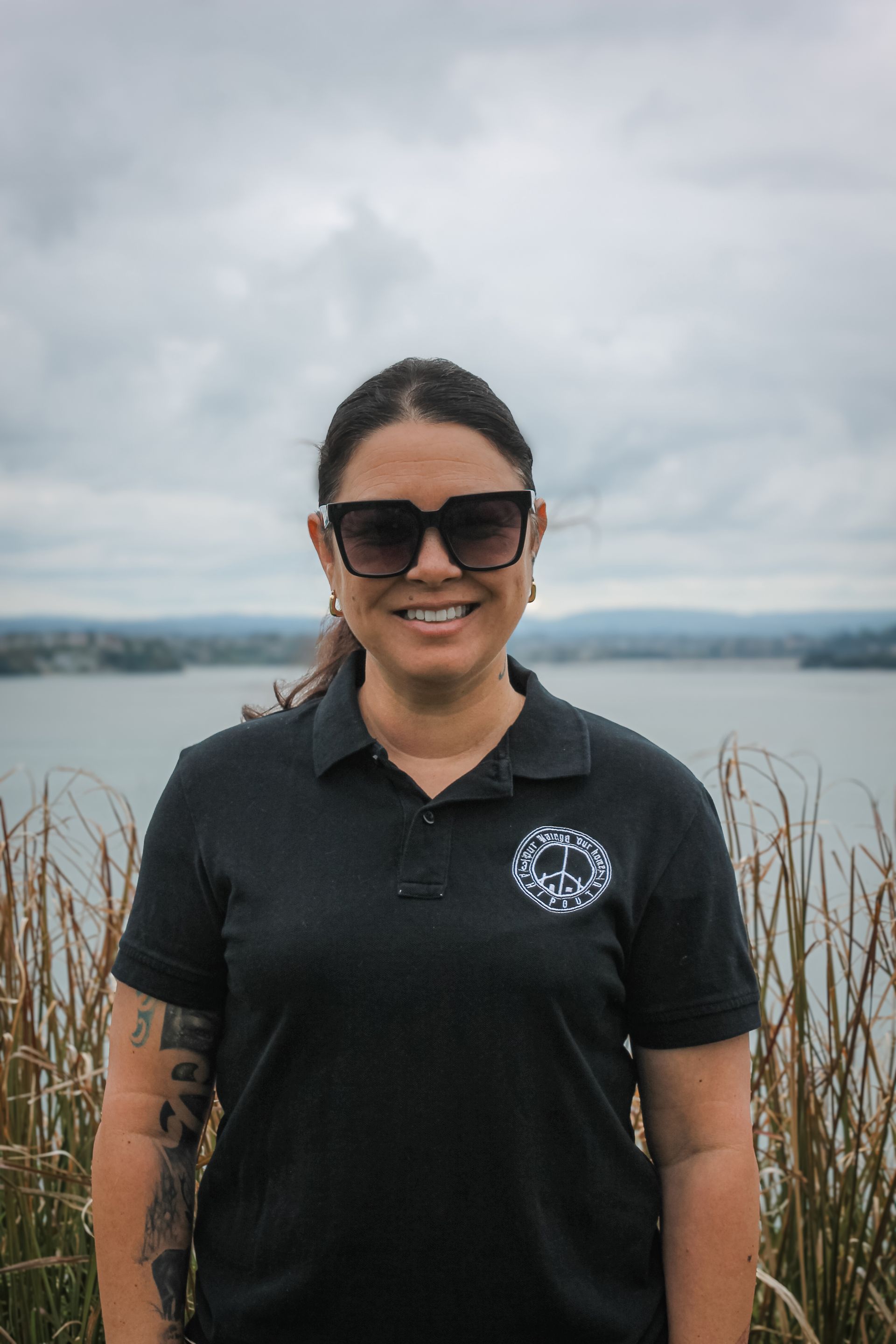 A woman wearing sunglasses and a black polo shirt is standing in front of a body of water.