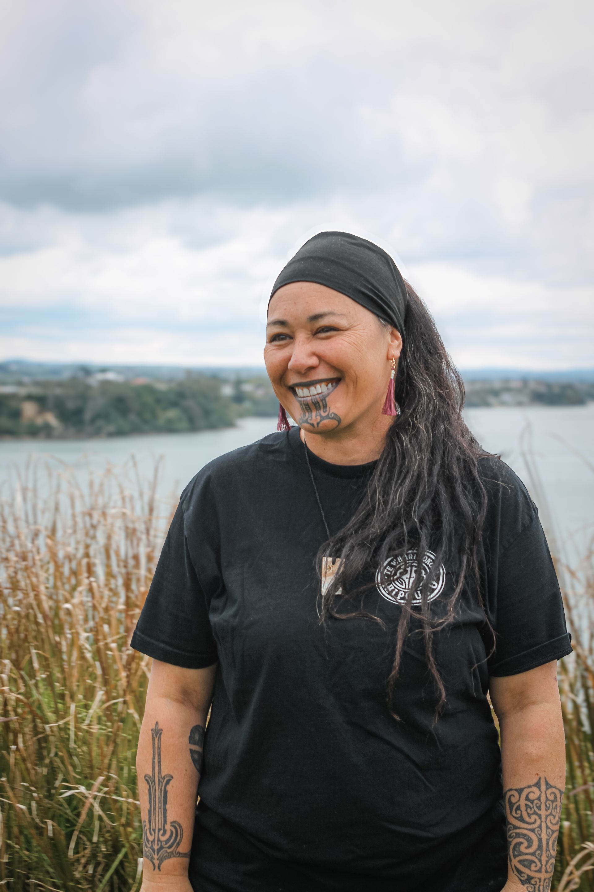 A woman with dreadlocks and tattoos is standing in a field next to a body of water.