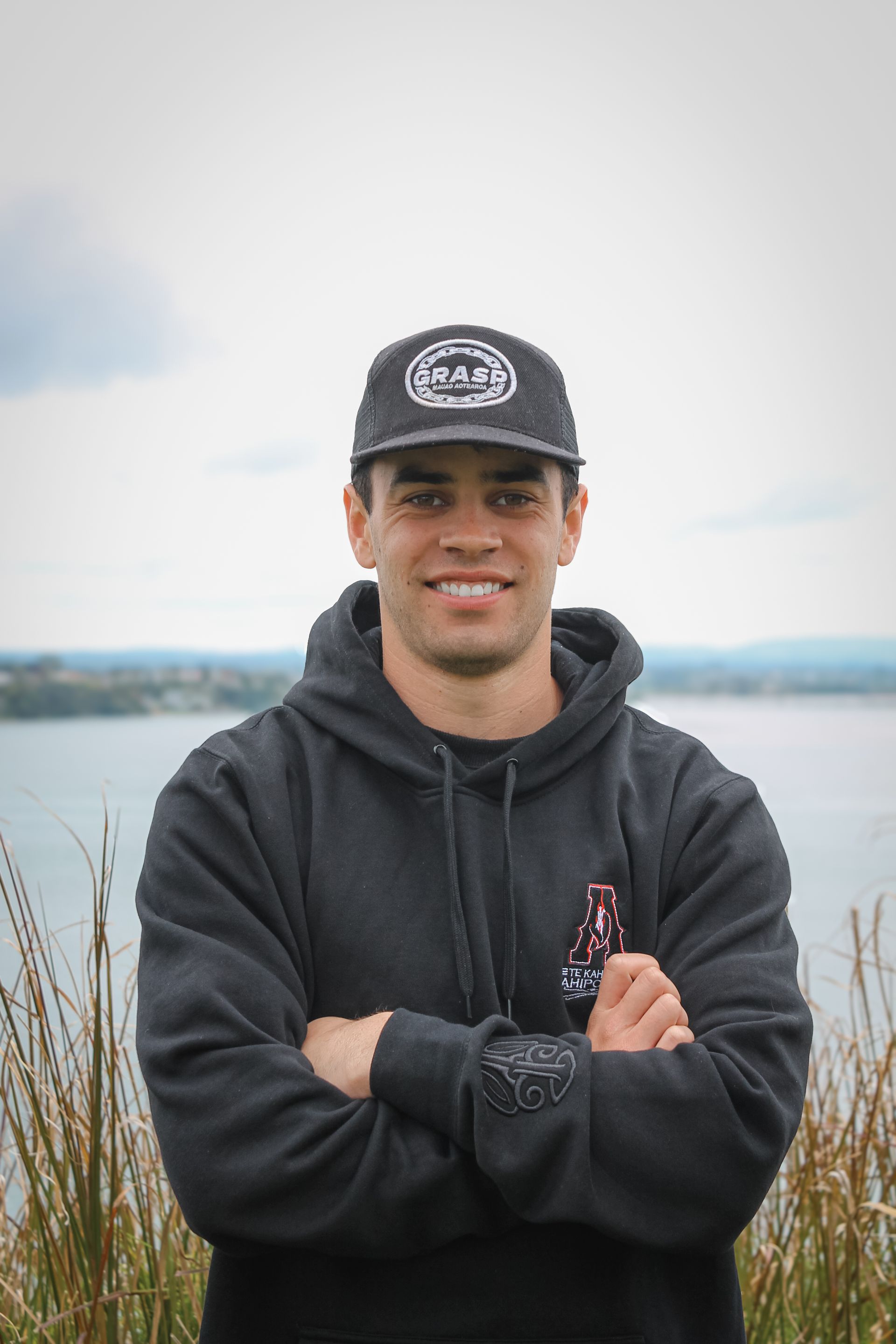 A man wearing a hat and a hoodie is standing with his arms crossed in front of a body of water.