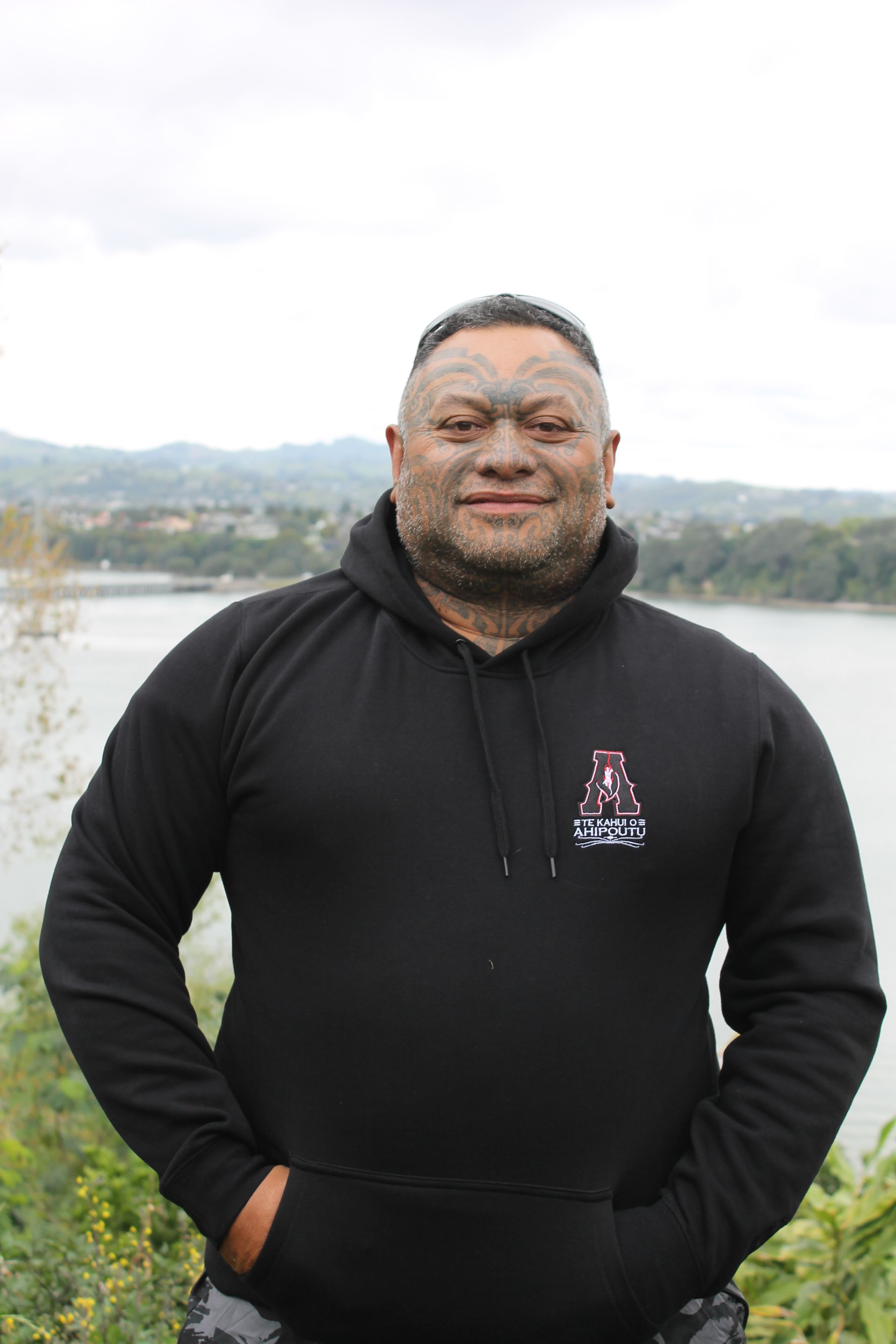 A man wearing a black hoodie is standing in front of a body of water.