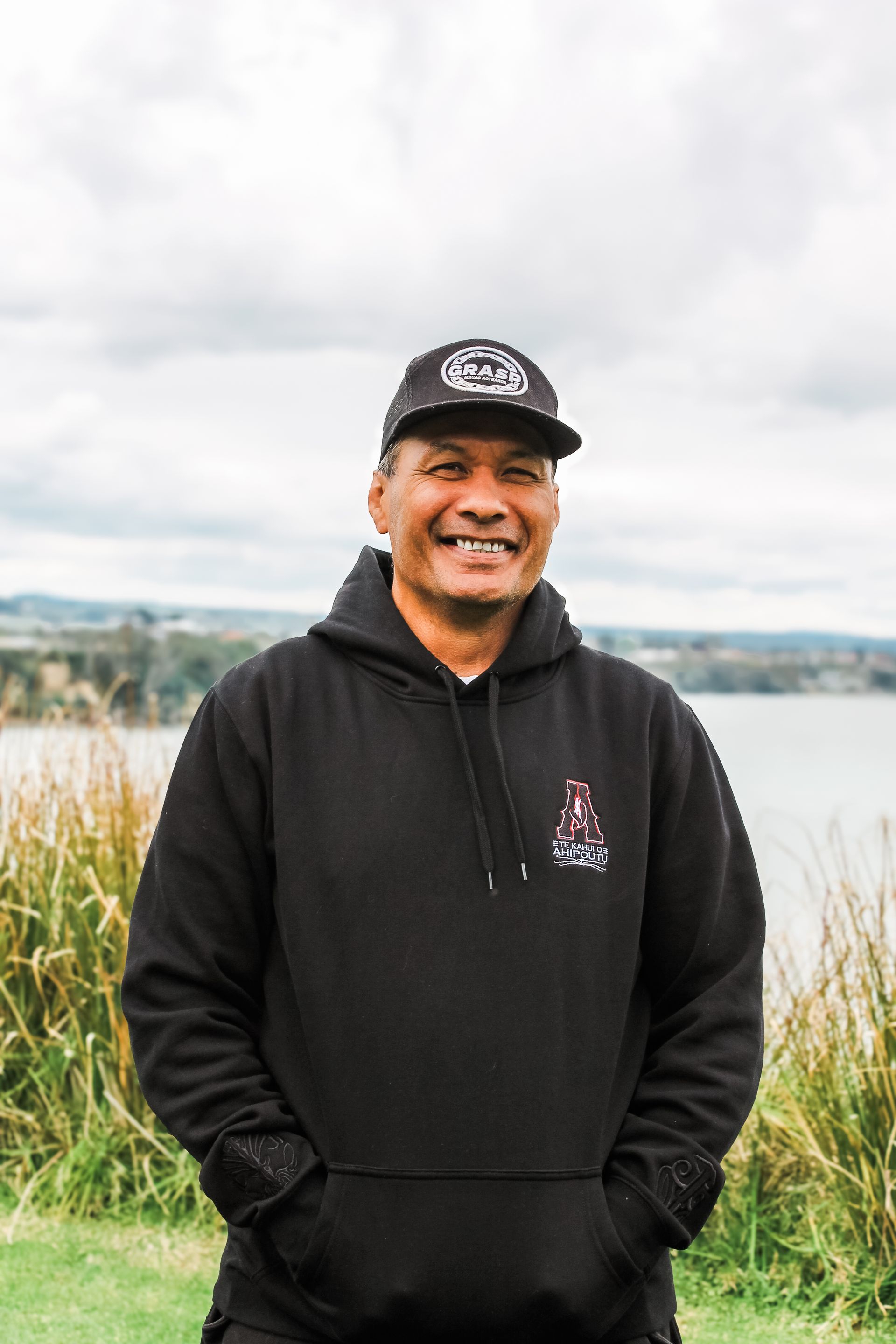 A man wearing a black hoodie and a hat is standing in front of a body of water.