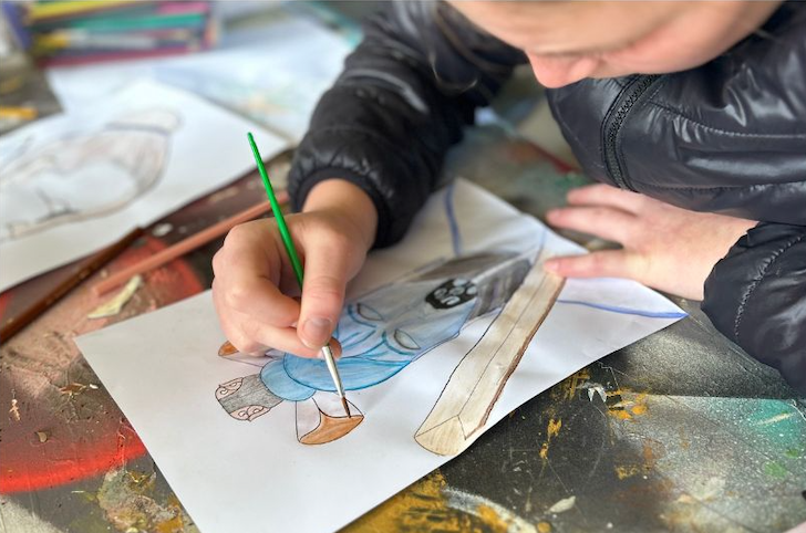 A young boy is painting a picture with a brush on a piece of paper.