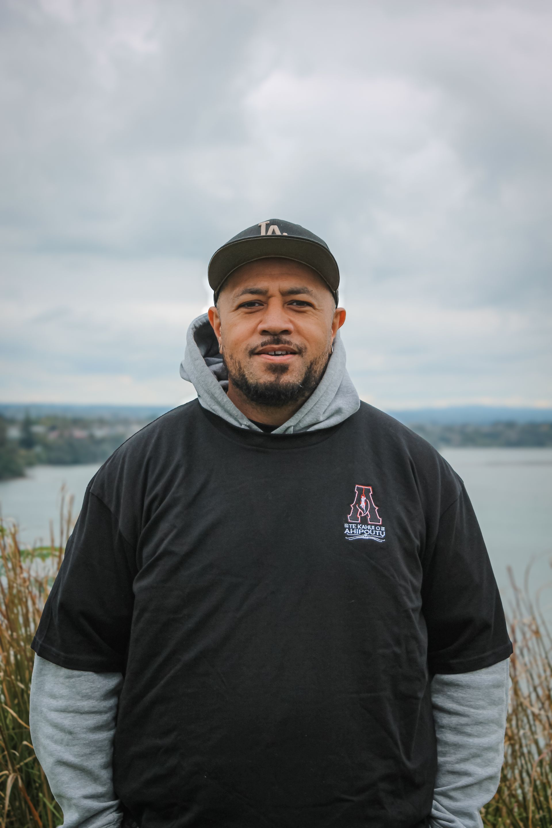 A man in a black shirt and hat is standing in front of a body of water.